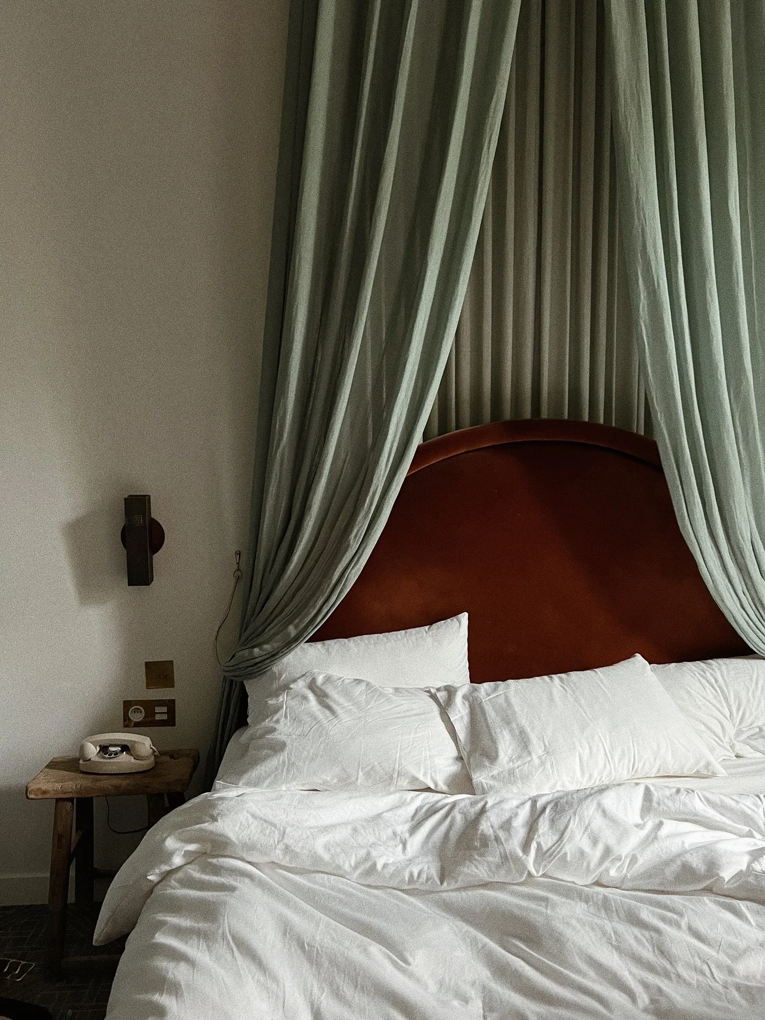 A neatly made bed with white sheets, pillows, a wooden nightstand with a vintage telephone, and a large, curved red headboard underneath green curtains in a hotel room.