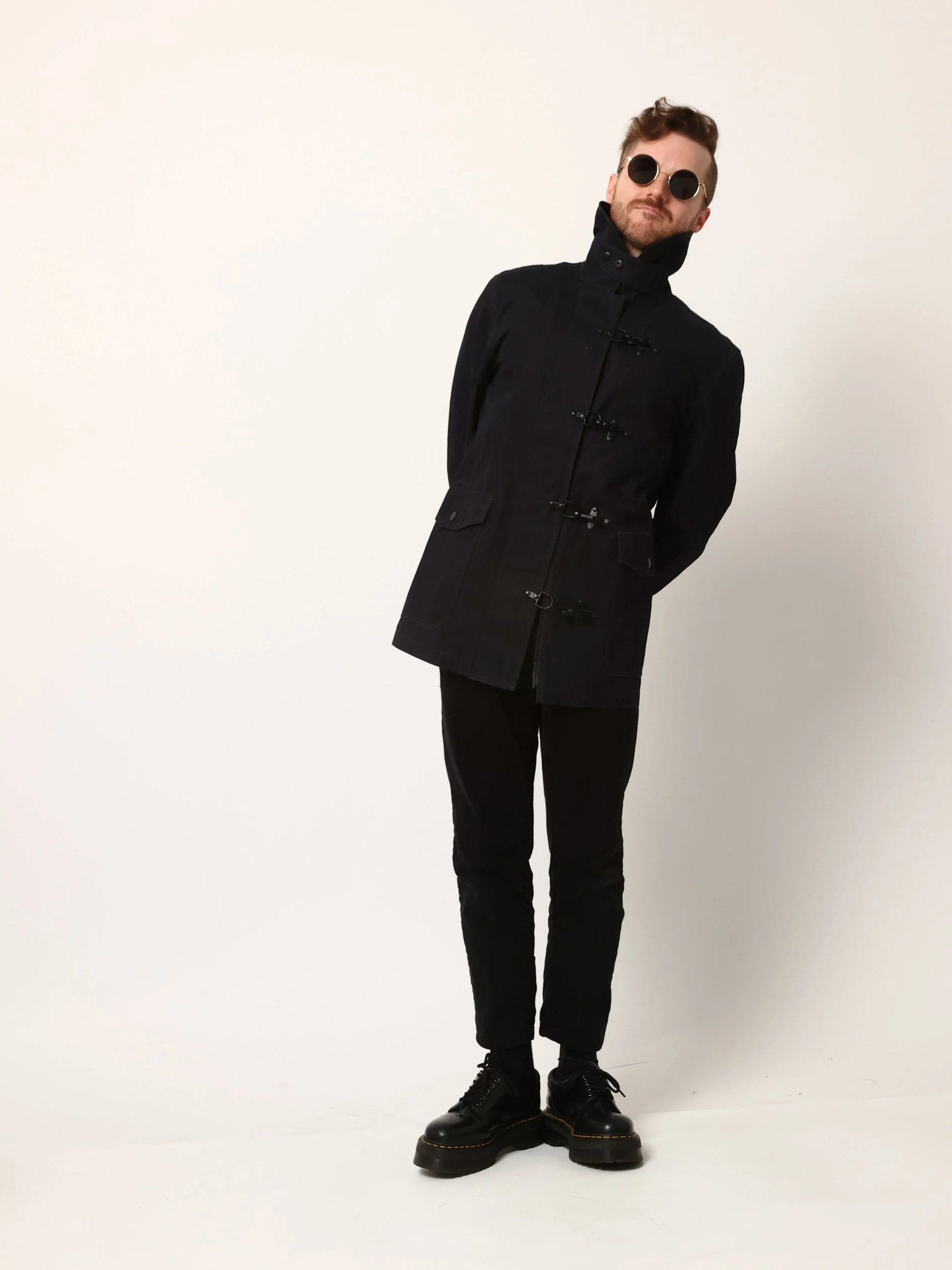 Mike Horton, leaning to the right in a black coat, black pants, and black shoes standing against a plain white background.