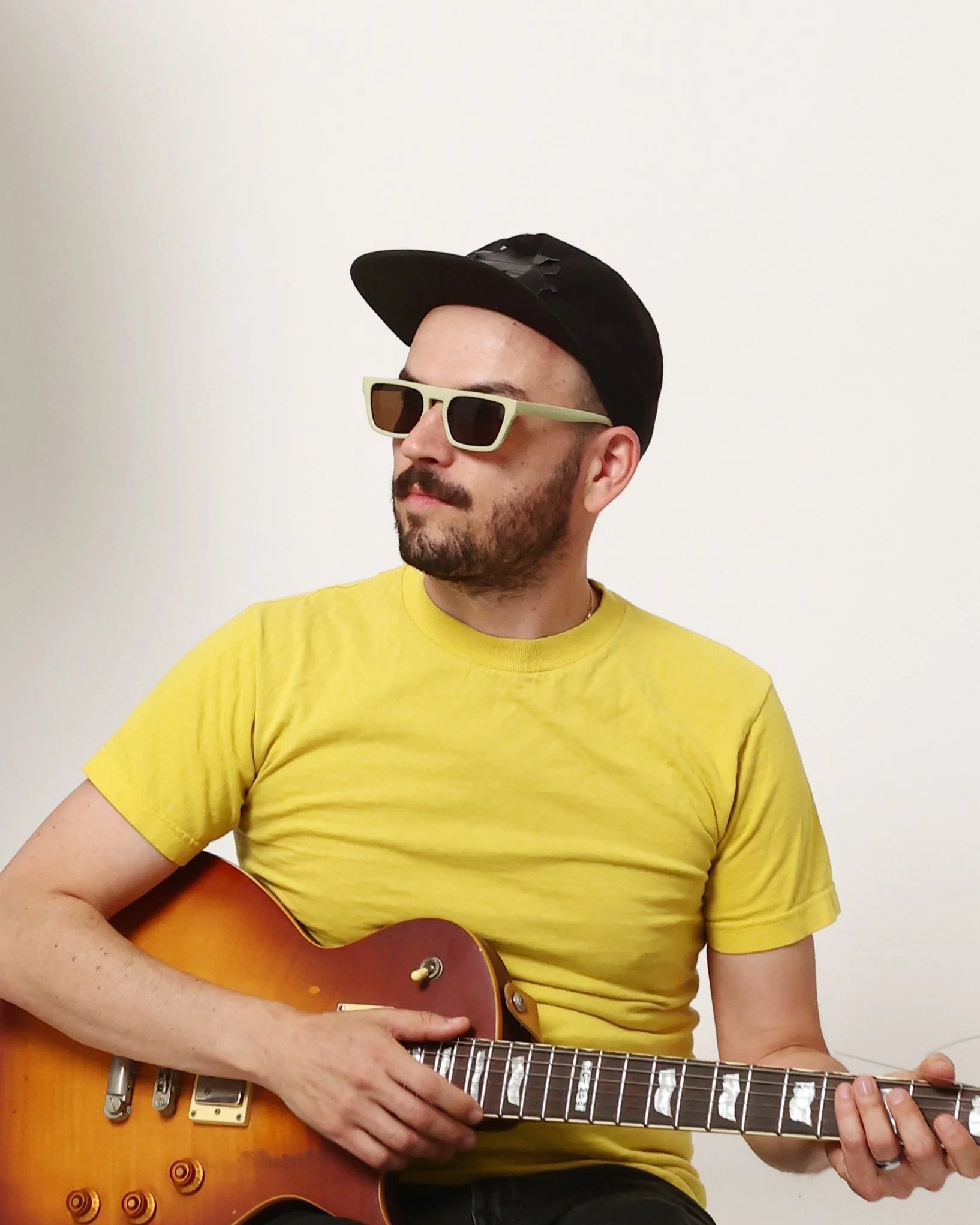 Eliot Murray wearing a yellow T-shirt, black backwards cap, and beige sunglasses, playing an acoustic guitar against a plain white background.