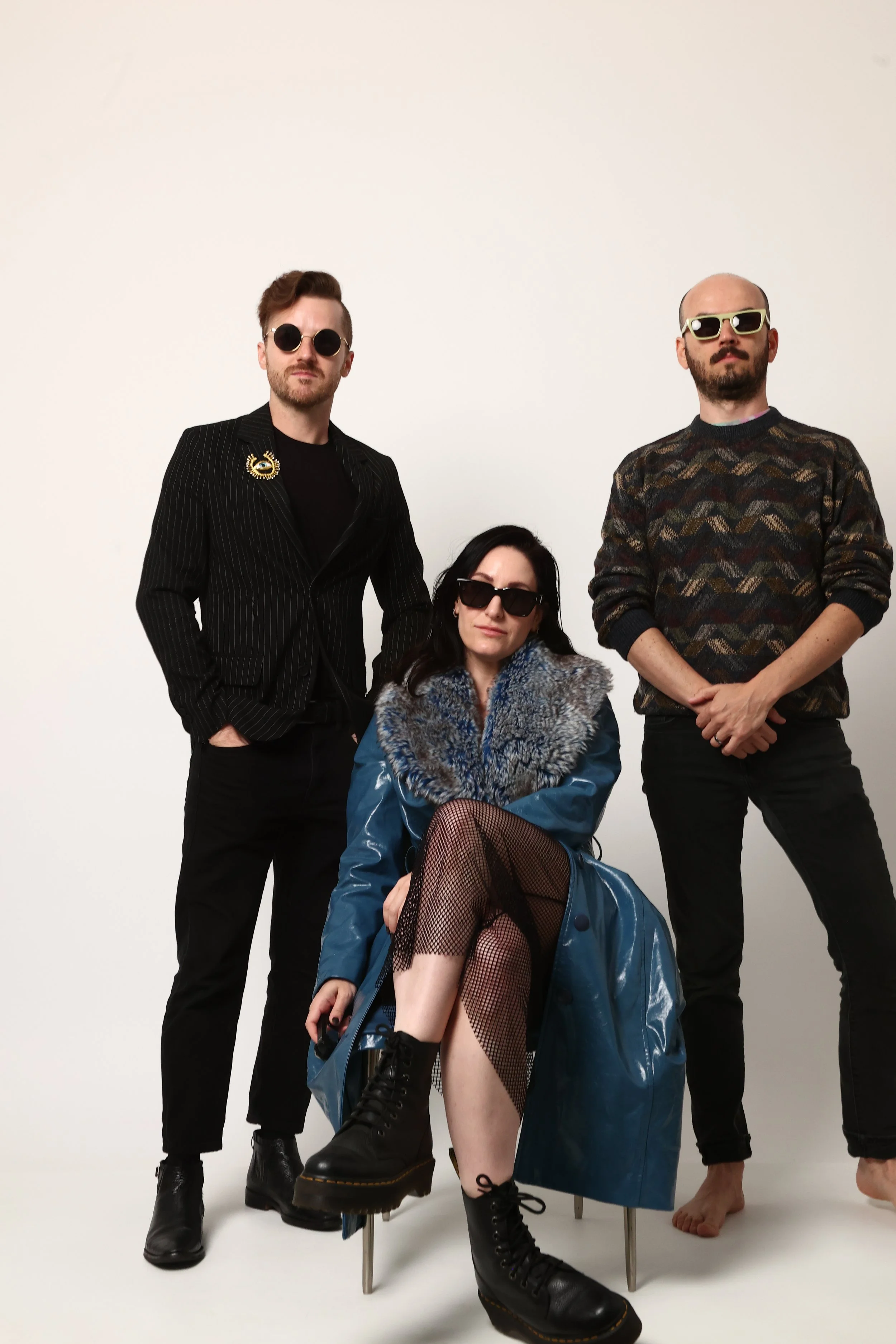 The three band members stylishly dressed on a white ground. One man stands with hands in pockets, another man stands with arms crossed, and a woman is seated, legs crossed, wearing a blue oversized coat and fishnet stockings.