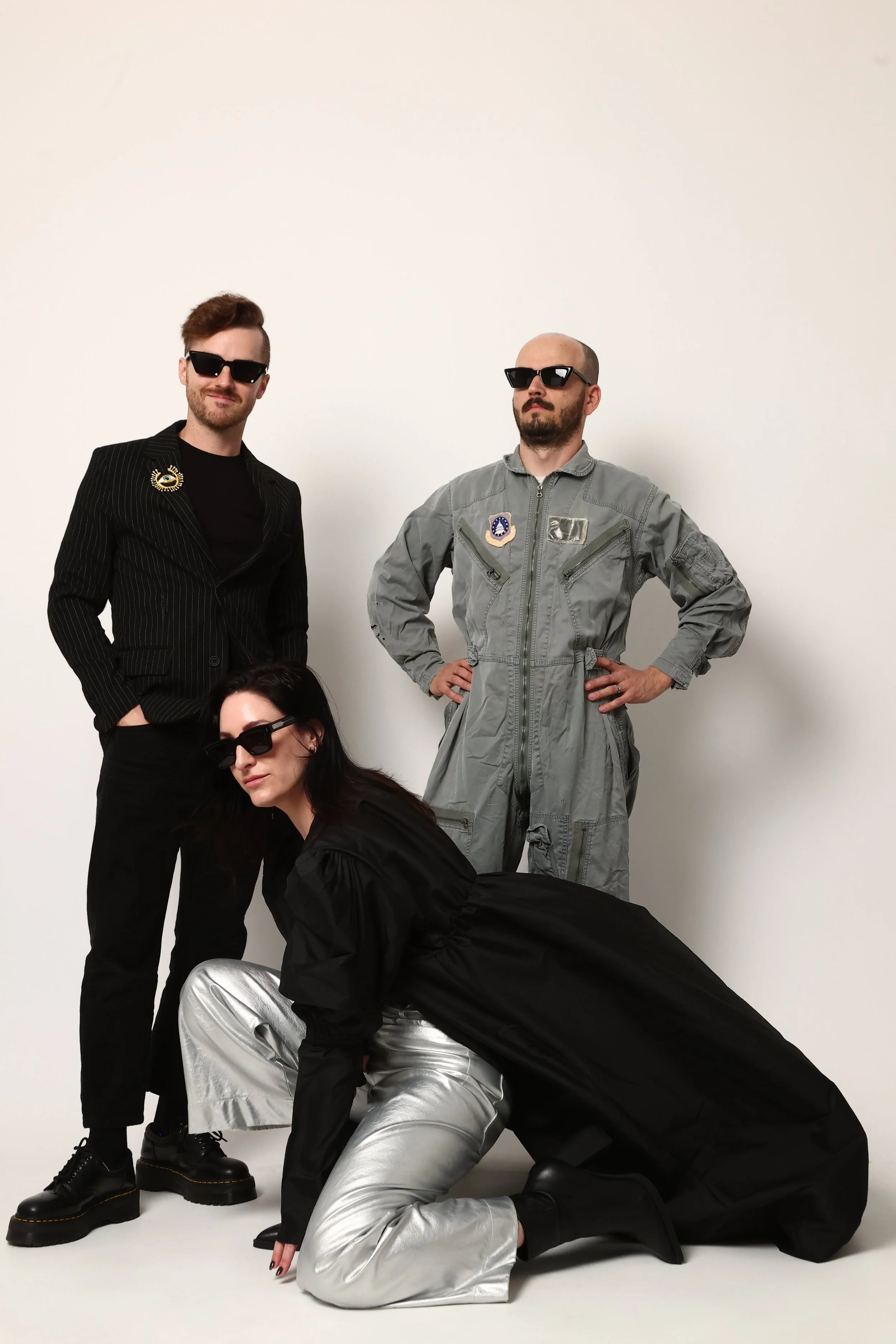 individuals in stylish, dark clothing and sunglasses pose against a plain, light-colored background. One person is squatting in front wearing silver pants, a black jacket, and dark footwear. Two others stand behind, one in a striped blazer with a brooch, and the other in a gray jumpsuit with patches. The fourth is partially visible at the bottom, wearing black clothing and silver pants.