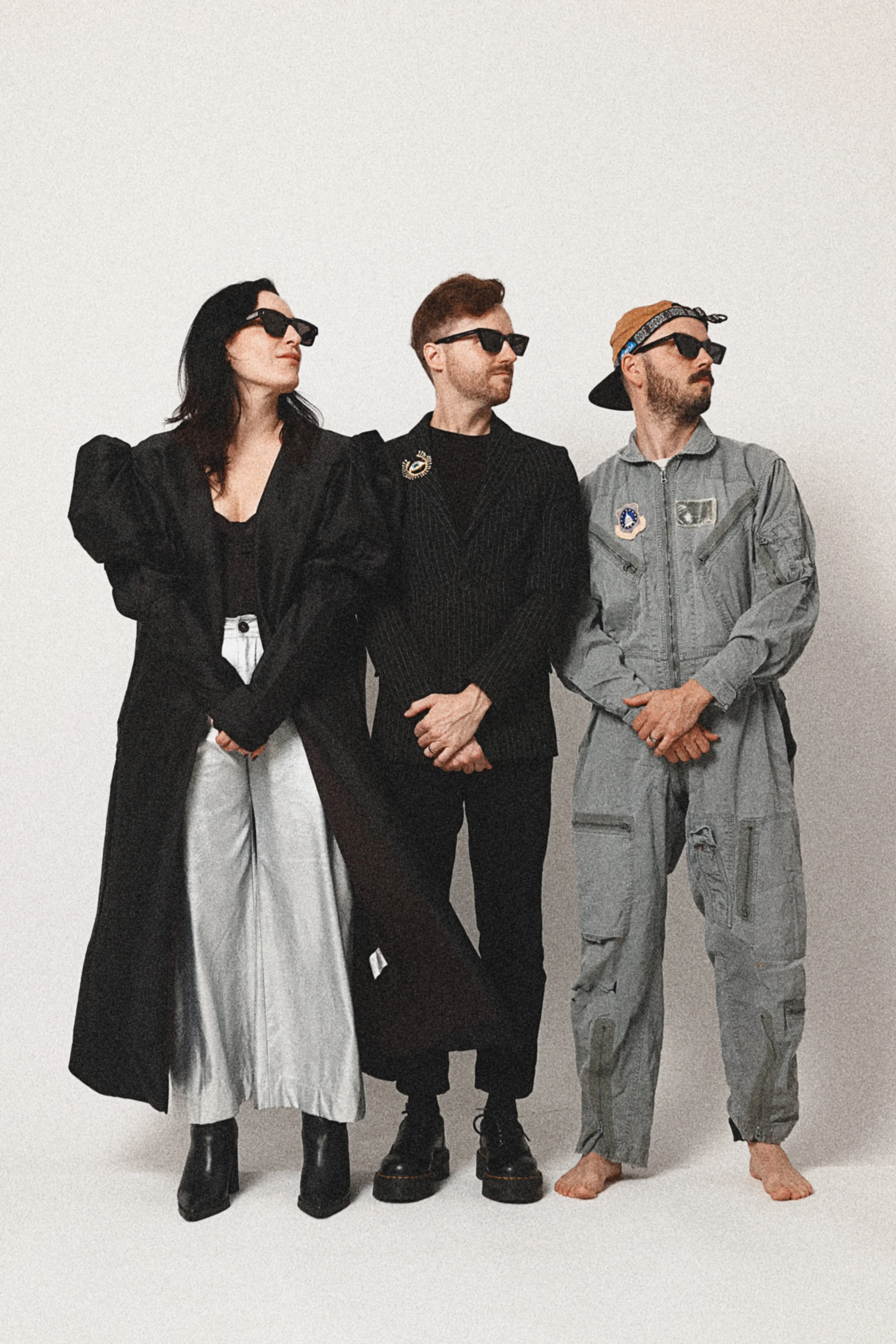 Three people standing side by side against a plain white background, wearing casual and streetwear style clothing, sunglasses, and accessories with faces 3/4 turned to the right.