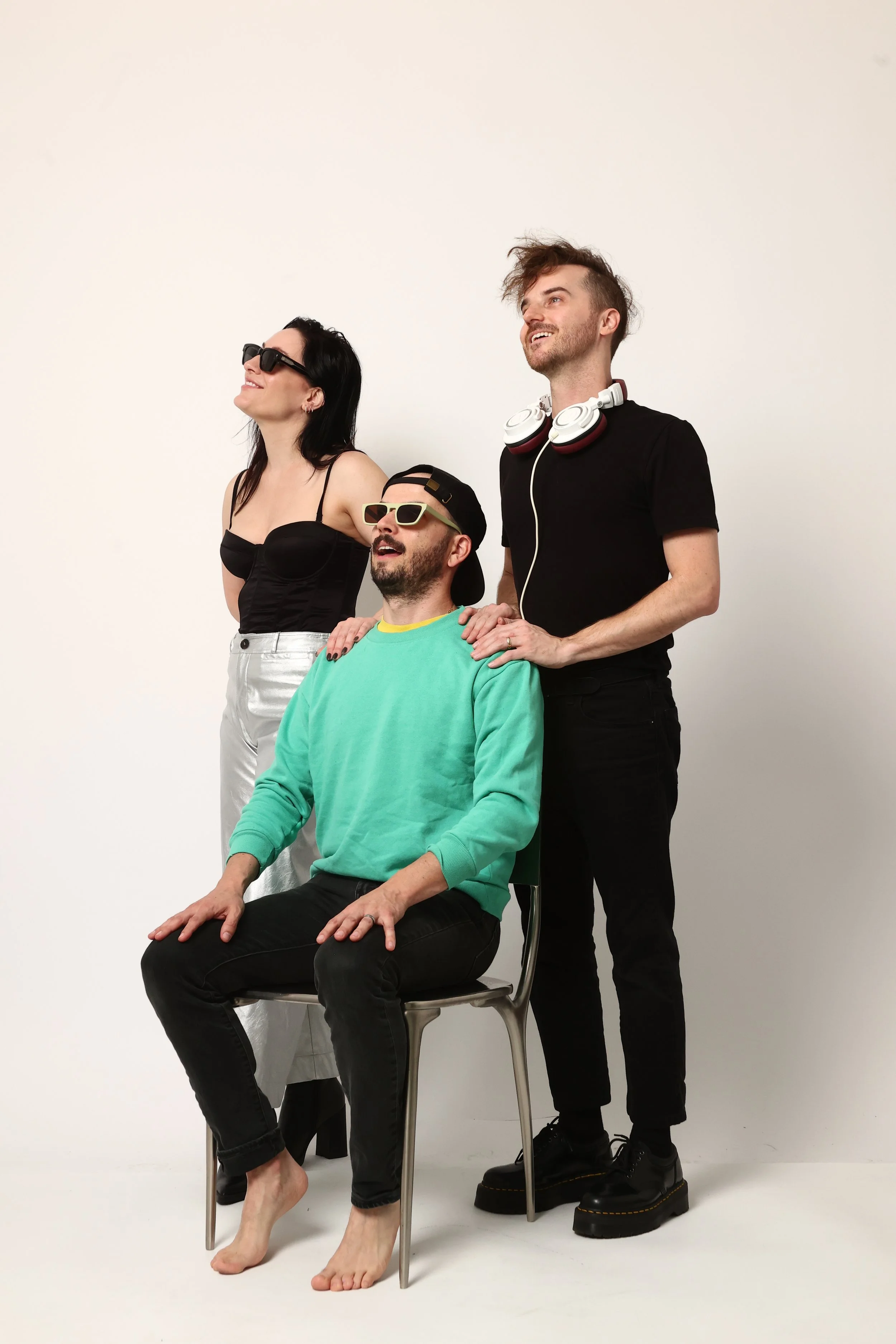 Three individuals posing: a woman in sunglasses and a black top standing to the left, a man in sunglasses with a cap seated in a chair in the middle, and a man with headphones and disheveled hair standing to the right with his hand on the seated man'