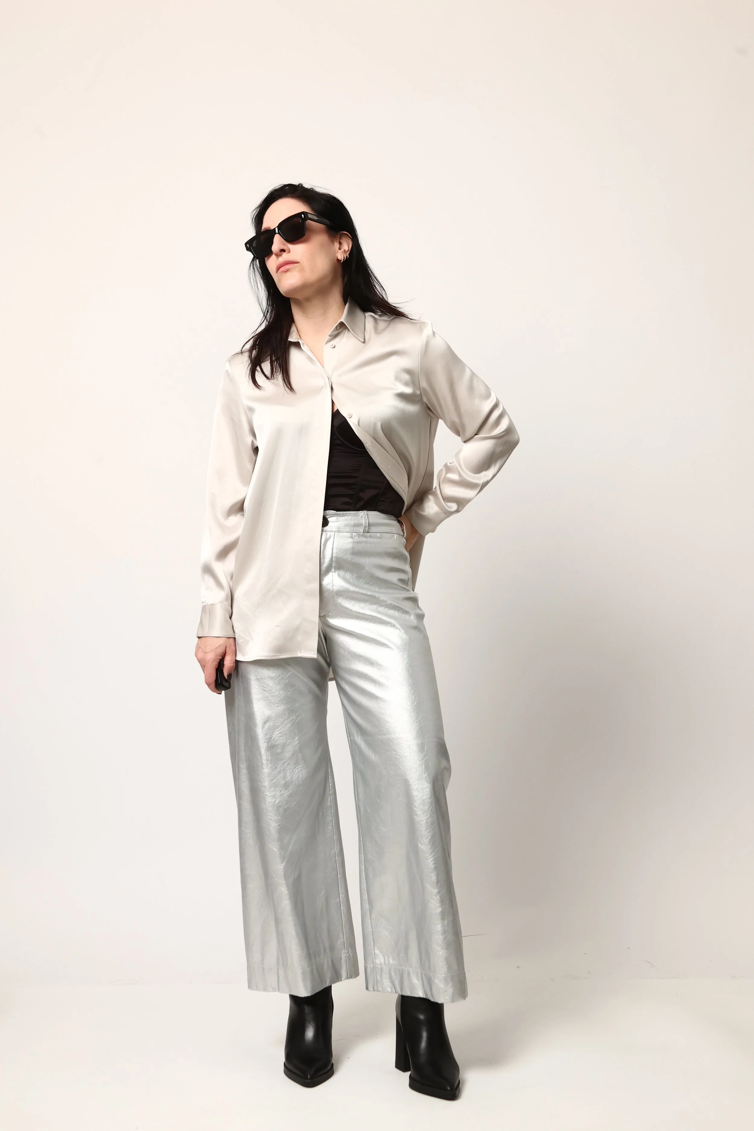 Nicole Cassese wearing black sunglasses, a satin cream blazer, a black top, shiny silver wide-leg pants, and black boots standing against a plain white background.