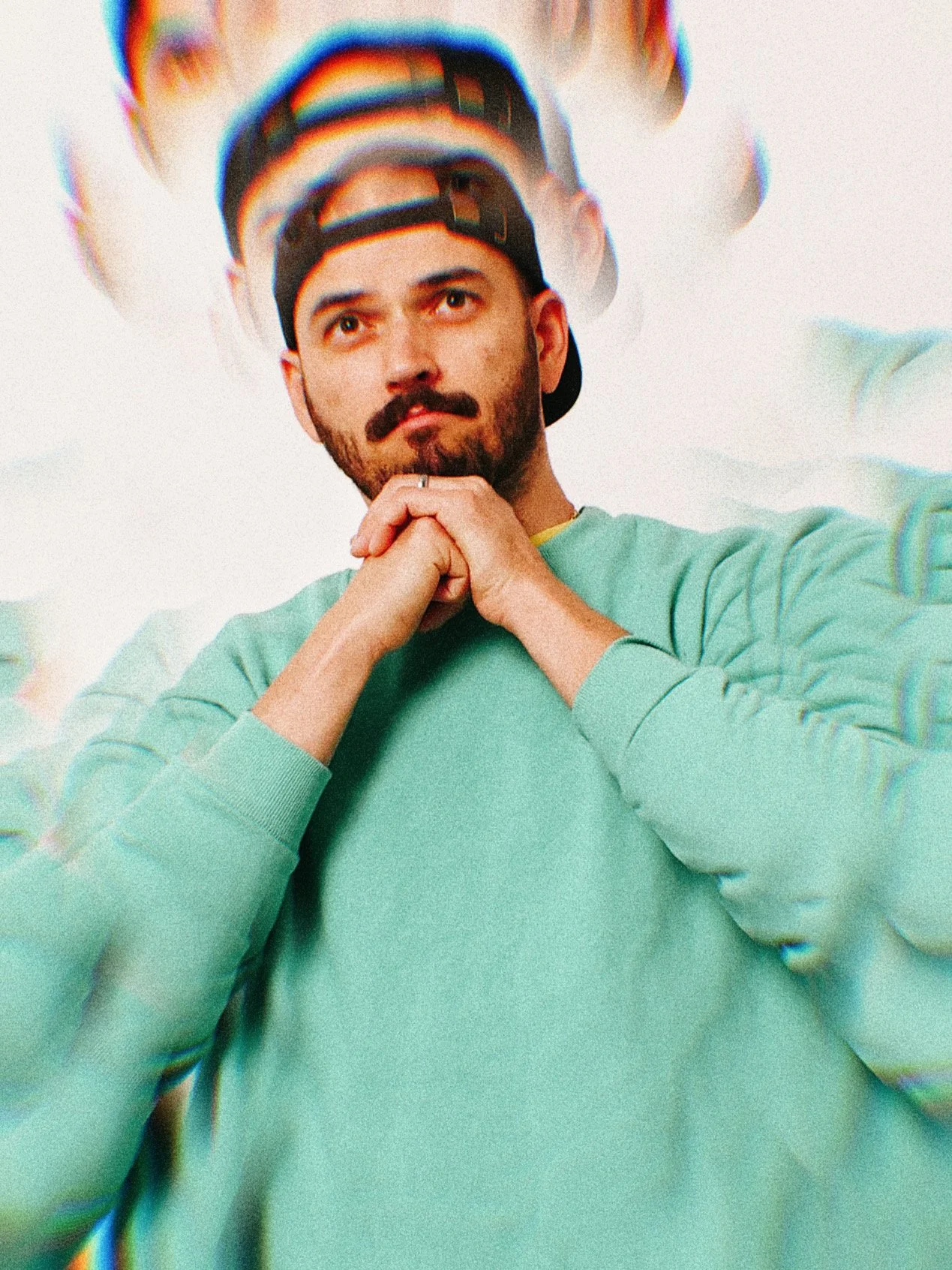 Eliot Murray, one of the lead musicians, featured with a beard, wearing a black baseball cap backwards and a mint green sweatshirt, is posing with his hands clasped under his chin. Digital distortion and multiple overlays create a glitch effect.