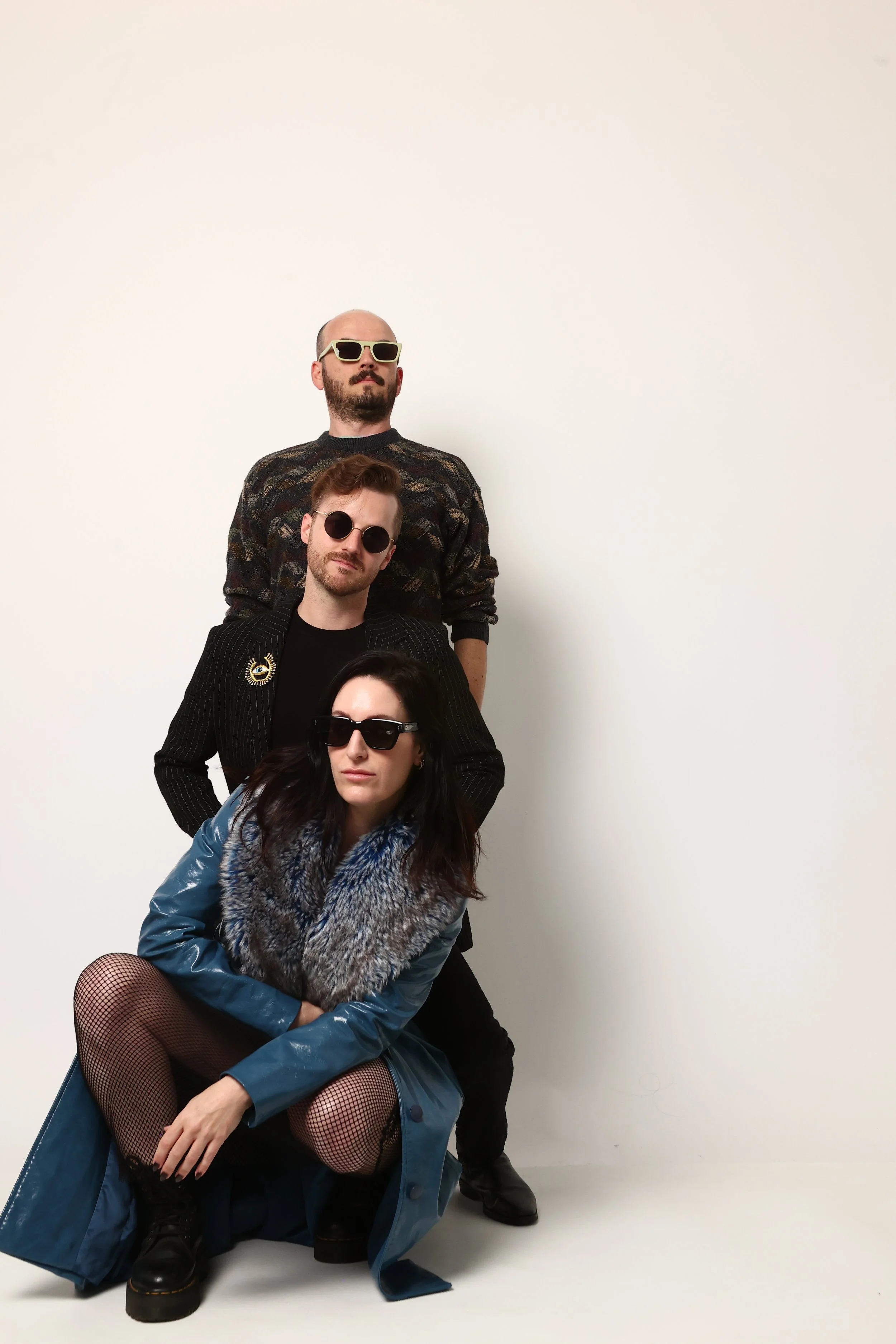 Group of the three band members posing stacked against a plain white background, wearing avant-garde fashion and sunglasses, with one model crouching in front and others standing successively behind.