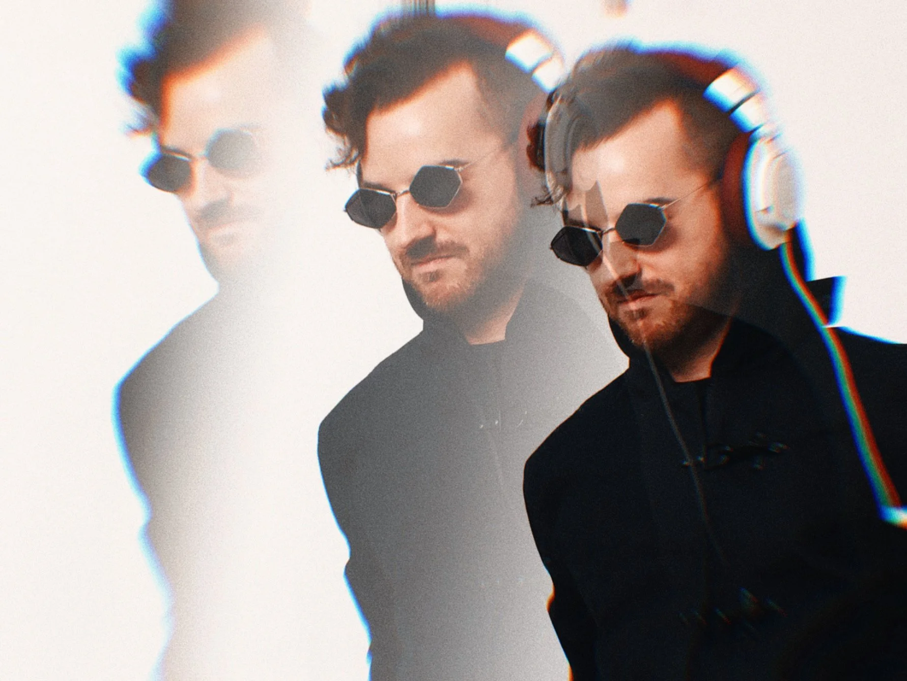 One man wearing a black jacket, sunglasses, and headphones posing for a photo with a white background. He is looking down or to the side with serious expressions.