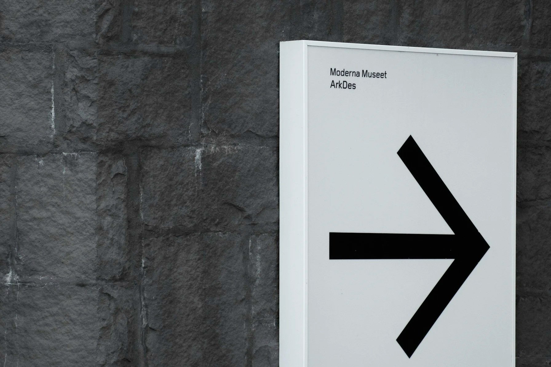 Sign pointing to the right with text 'Moderna Museet ArkDes' in the top left corner, mounted on a gray brick wall.