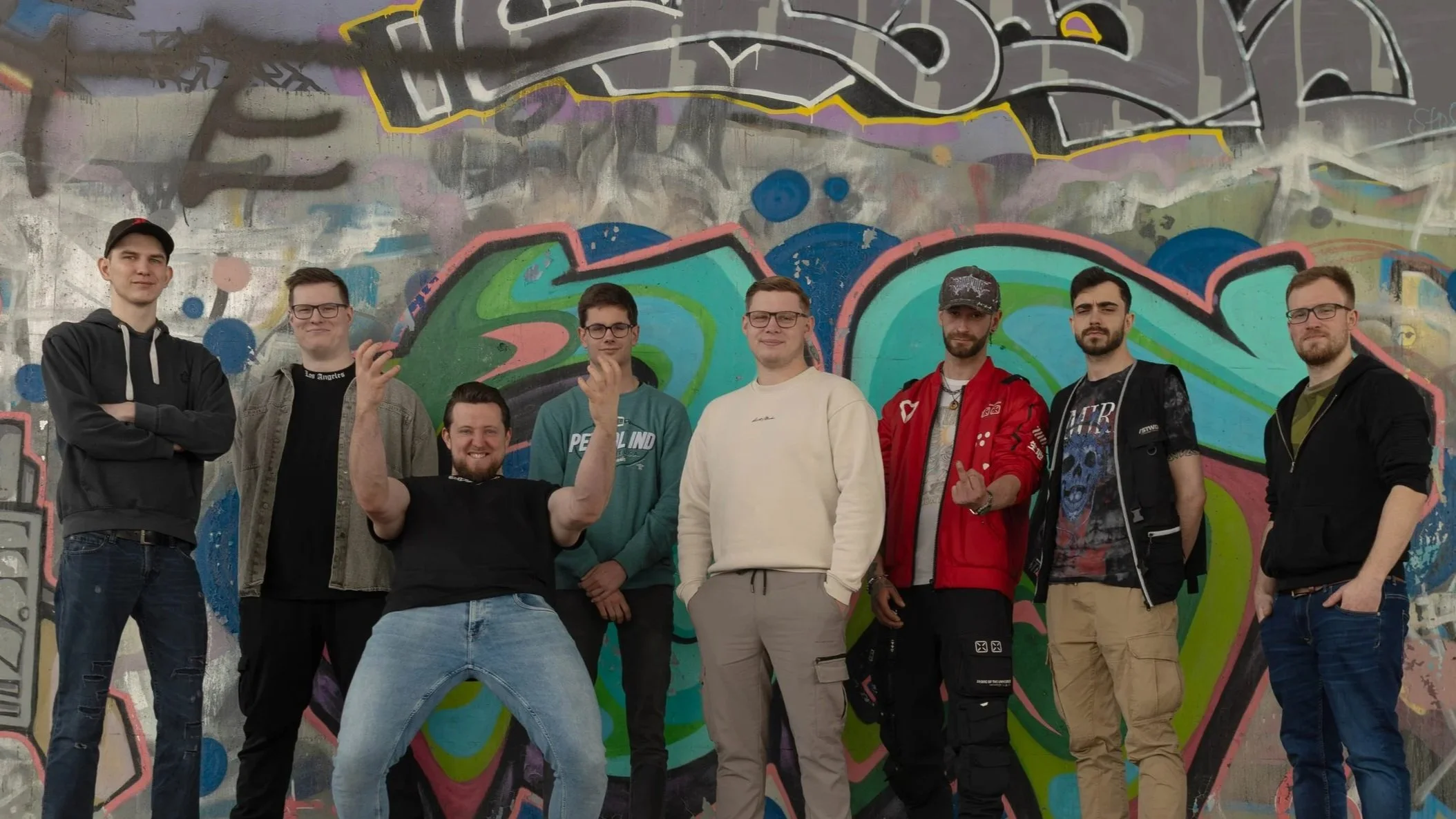 Group of eight young men standing in front of colorful graffiti wall, some smiling, some with casual poses.