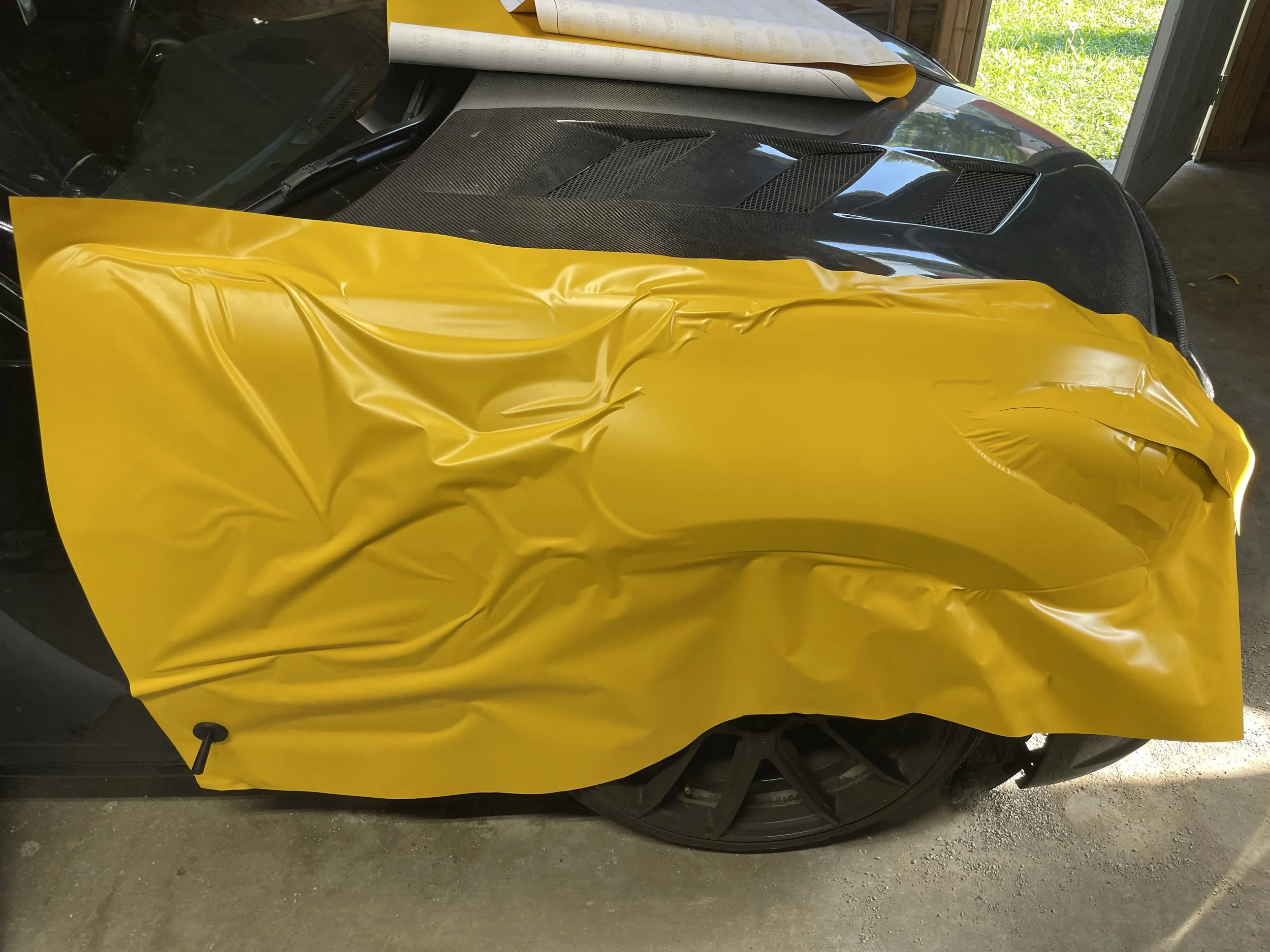 A portion of a black sports car with a yellow vinyl wrap partially applied to the front fender and hood, in a garage with a grassy outdoor view.