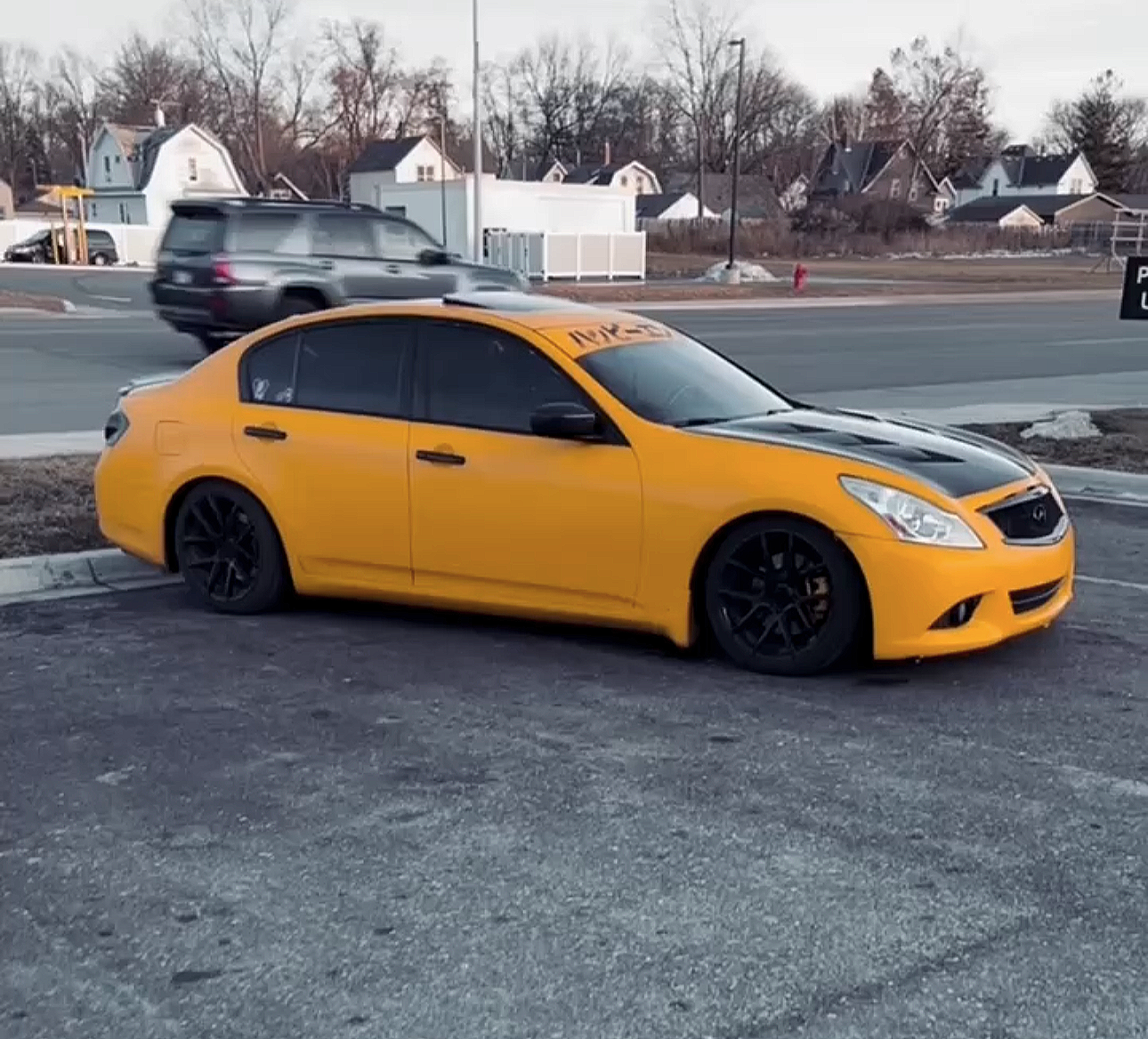 A yellow sedan with black hood and blacked-out wheels parked on an asphalt lot, with a road and houses in the background.