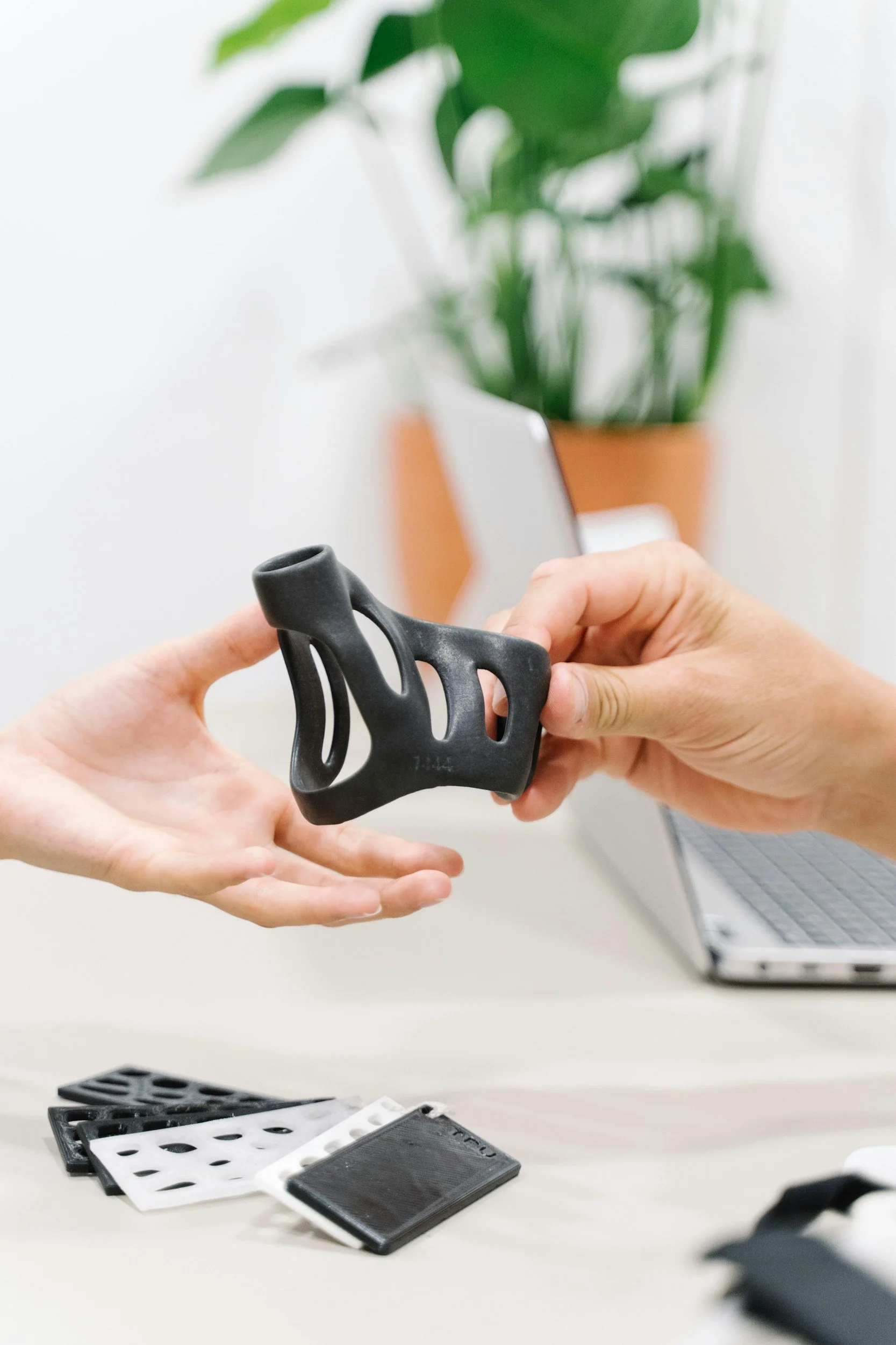 Two hands exchanging a black 3D printed object over a table with color samples, with a laptop and a plant in the background.
