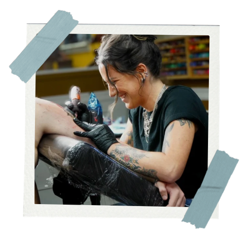 A woman with tattoos and black gloves is tattooing a person's arm in a tattoo shop, smiling and focusing on her work.