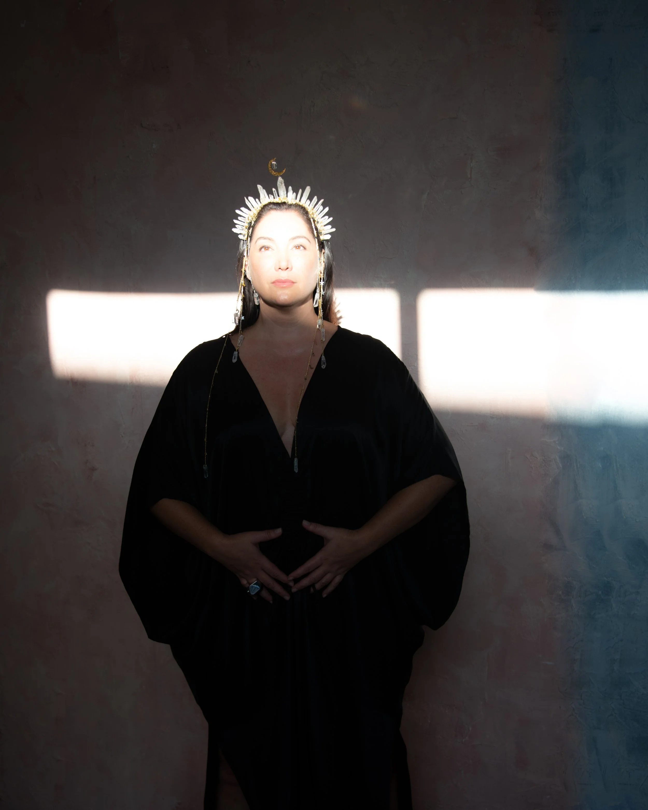 A woman with a radiant sunlit face wearing a black robe and an ornate headdress, standing against a textured wall with sunlight creating a rectangular pattern behind her.