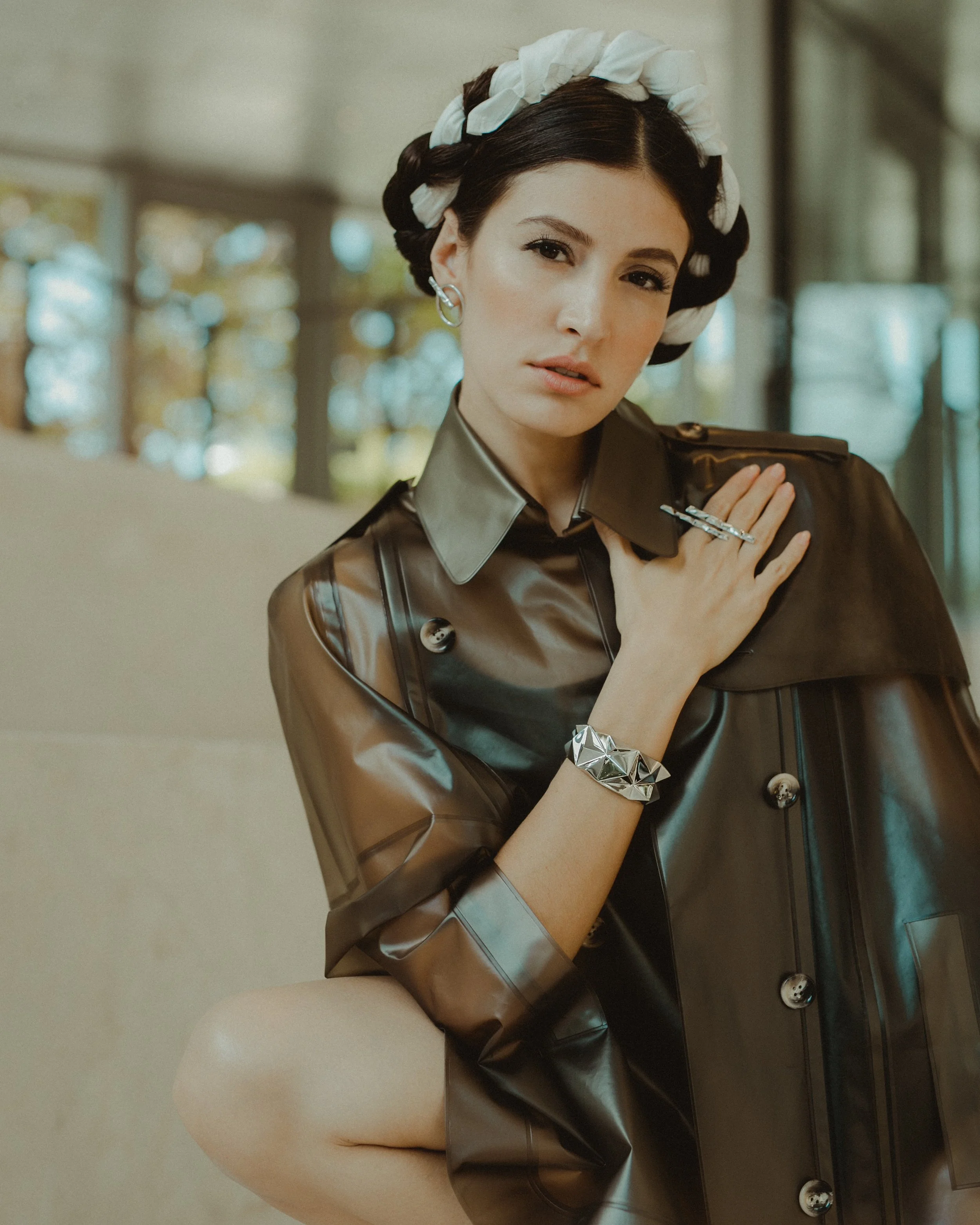 A woman with dark hair styled in vintage waves, wearing a large white headband, and dressed in a shiny brown leather jacket, sitting on a beige sofa in a modern indoor setting with large windows and blurred outdoor scenery. She has jewelry including 