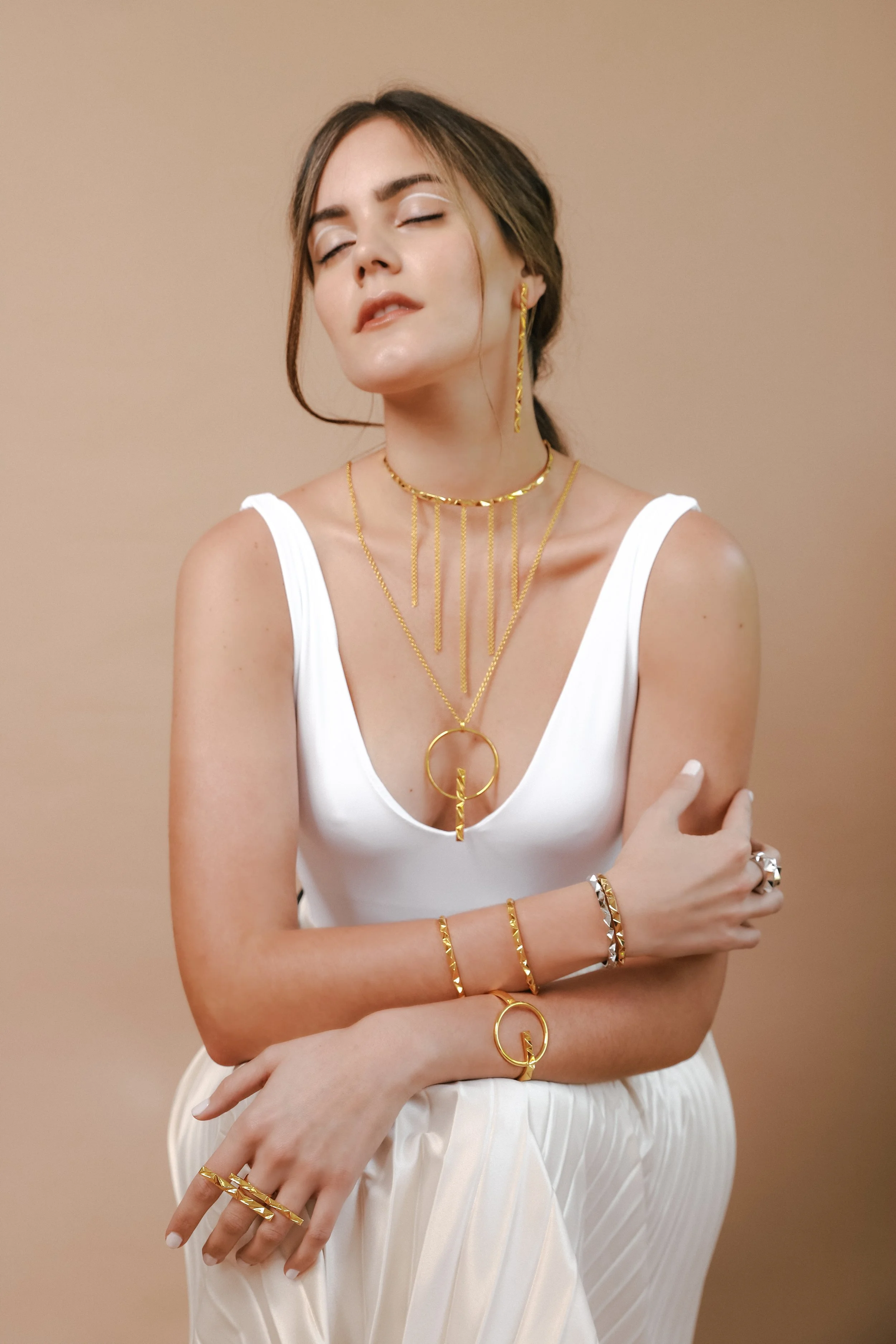 A woman wearing a white sleeveless top and light-colored pleated pants adorned with multiple gold jewelry pieces, including necklaces, bracelets, rings, and earrings, standing against a beige background with her eyes closed.