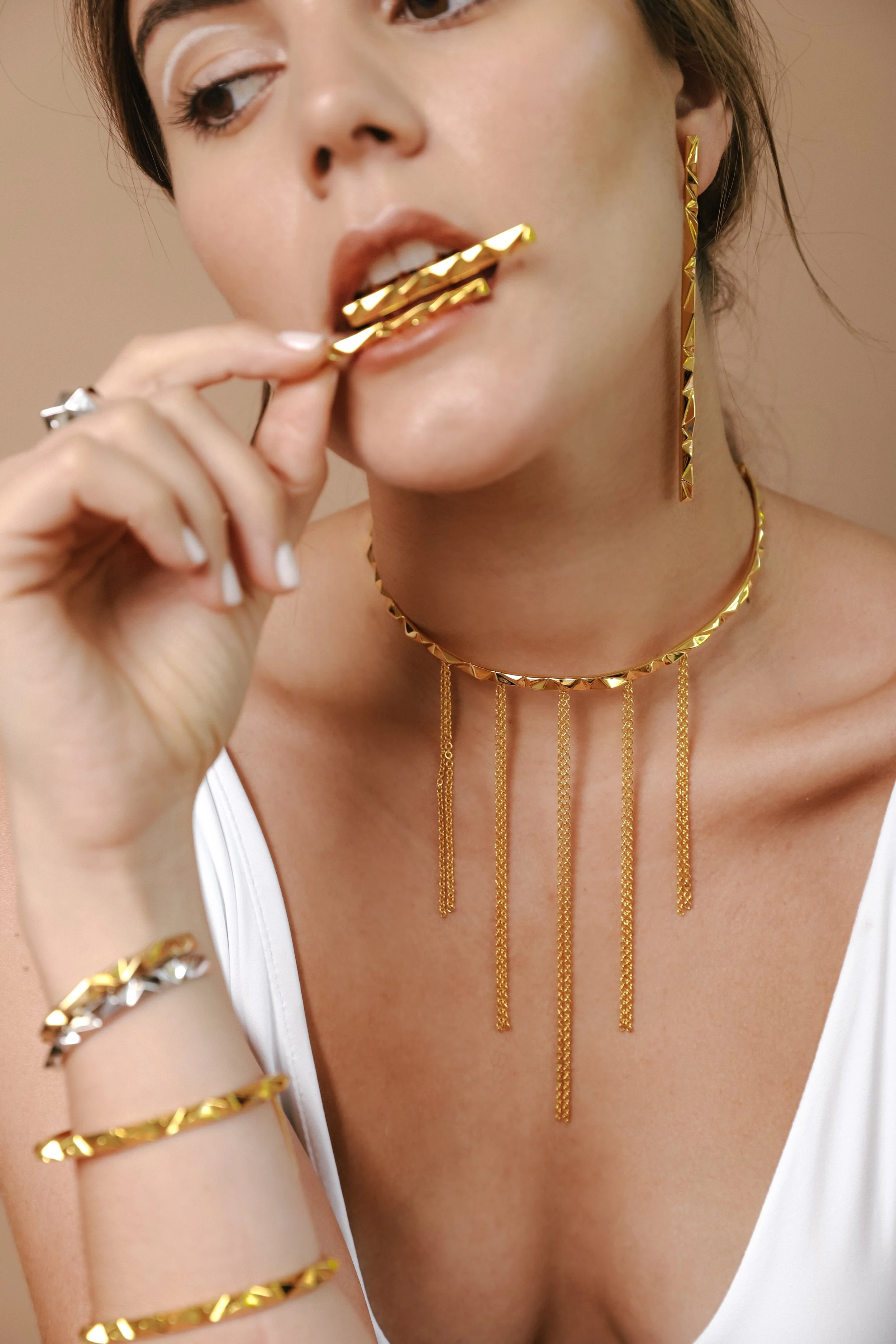 A woman with brown hair and fair skin is wearing gold jewelry, including a necklace, earrings, rings, and bracelets. She is holding a gold bar to her lips, with her mouth slightly open.