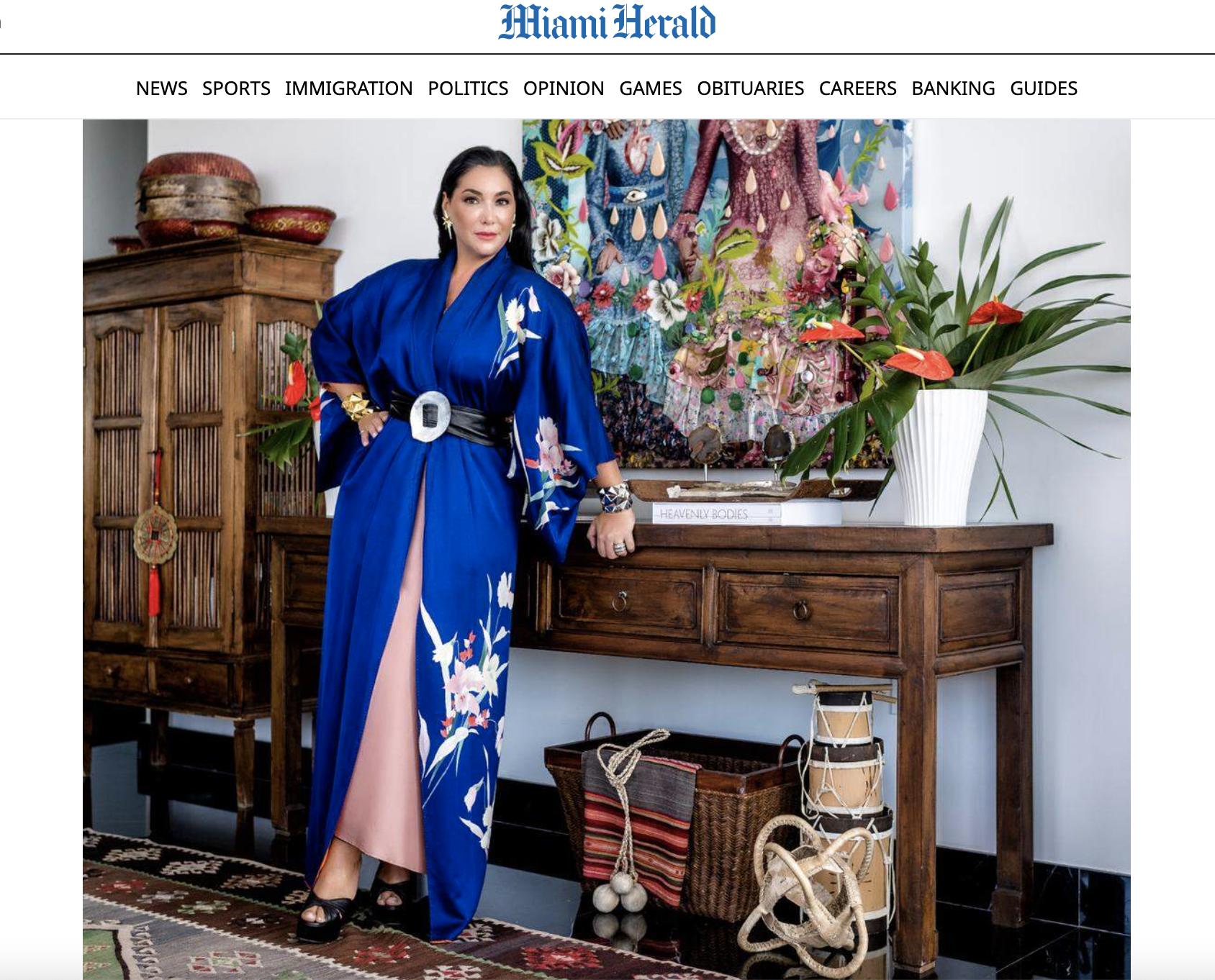 Woman in a blue floral kimono-style dress standing indoors next to a wooden cabinet and a side table with a large white vase of red and green plants, colorful textile art hanging on a white wall.