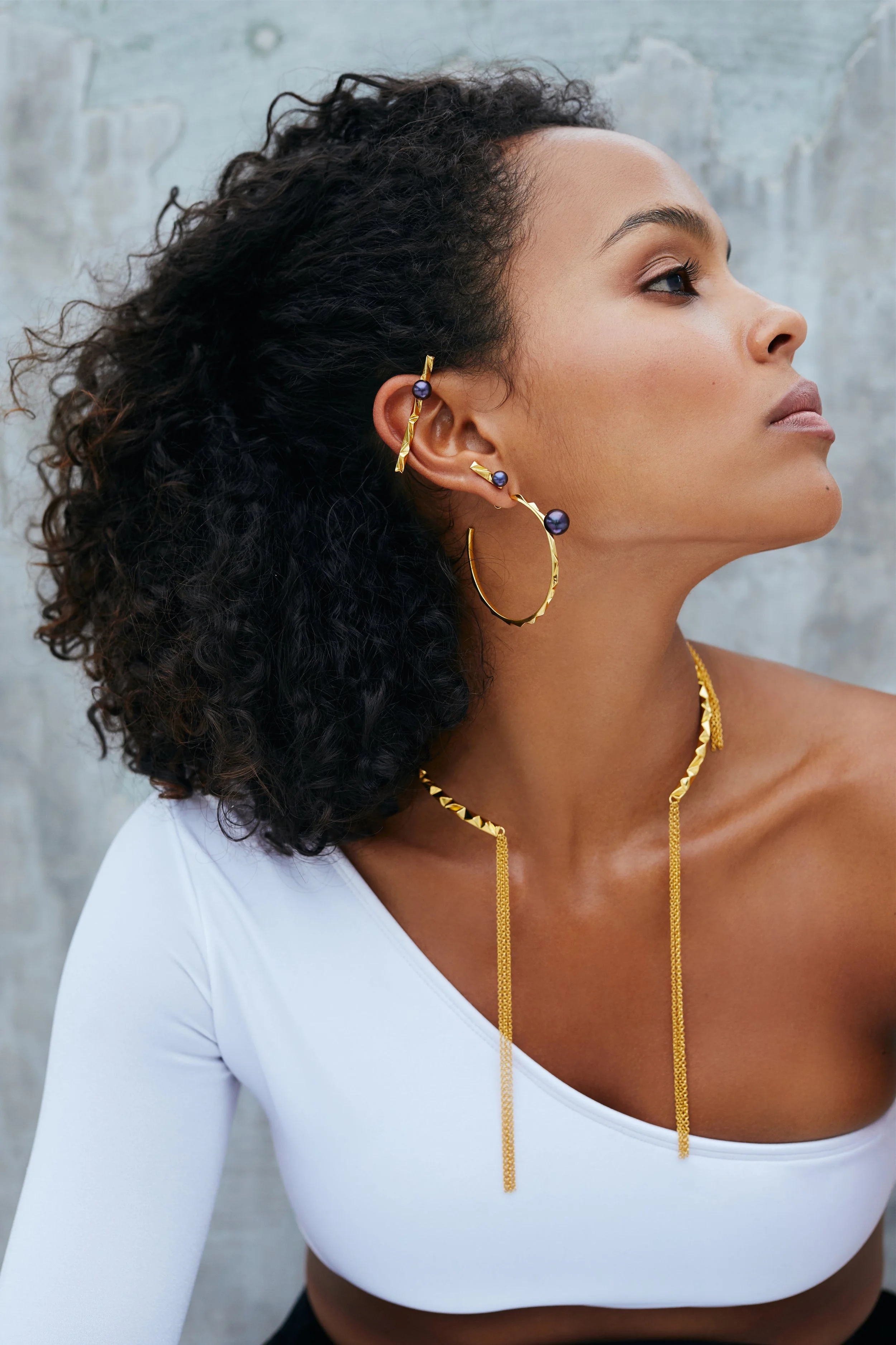 A woman with curly dark hair wearing gold jewelry including earrings, a necklace, and a bracelet, against a gray concrete background.