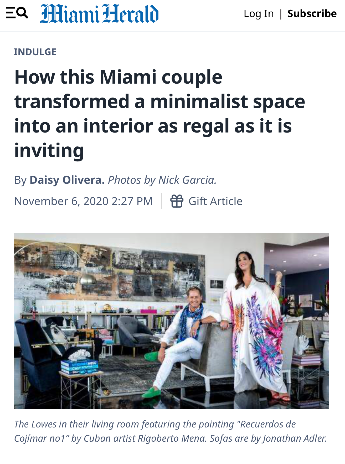 A photo of a couple inside a modern living room. The man is sitting on an armchair, smiling, wearing a white shirt, white pants, and green shoes. The woman is standing beside him, smiling, wearing a colorful, floral-detailed dress. The room features 