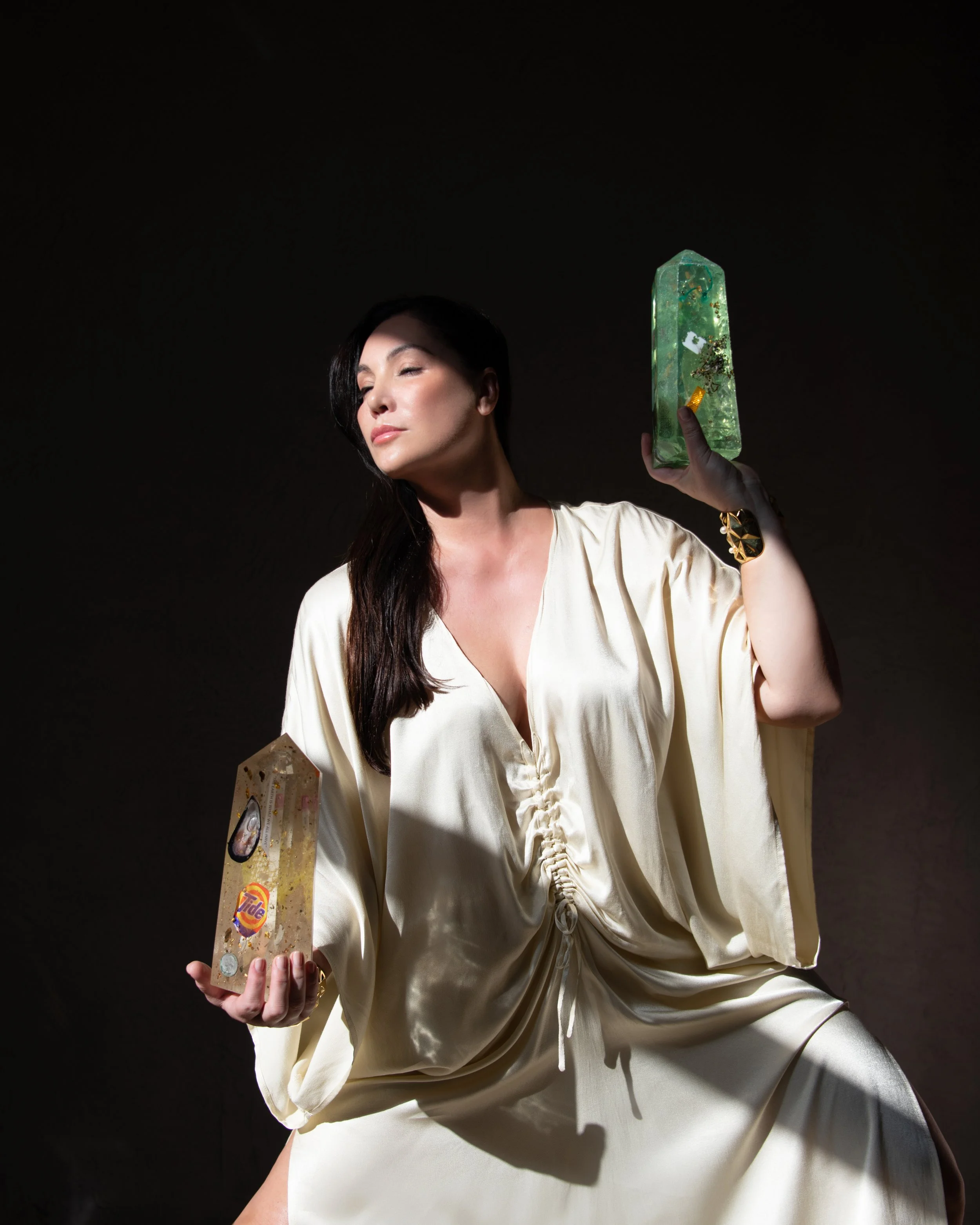 A woman with long dark hair in a cream satin outfit holding two soap bars, one with a TIDE logo and the other a green decorative soap, against a dark background.