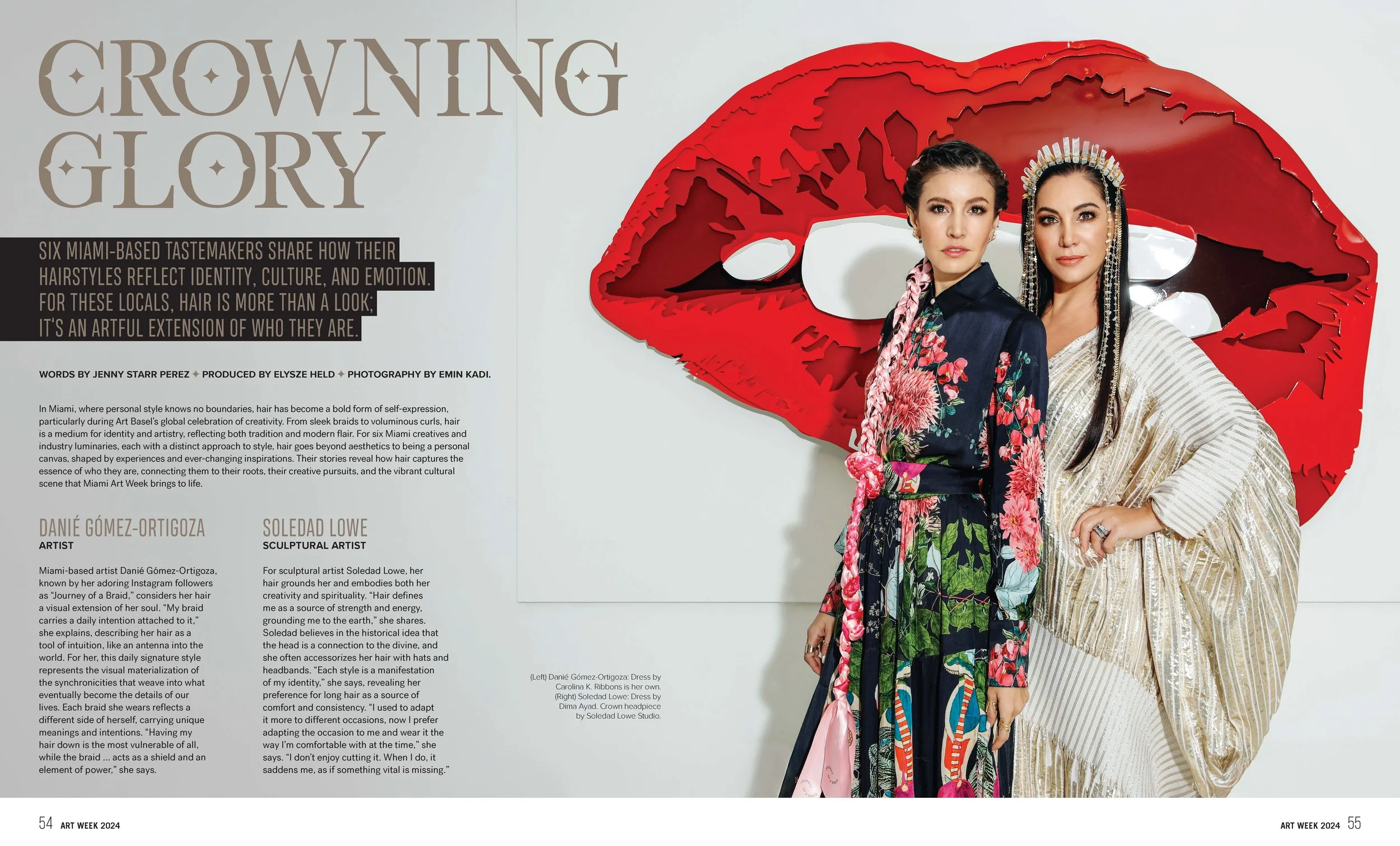 Two women standing in front of a large red lipstick-shaped art installation. One wears a dark floral robe with pink accents, the other a shiny gold gown with a headpiece. The background features graphic lips and text about hair inspiration and cultur