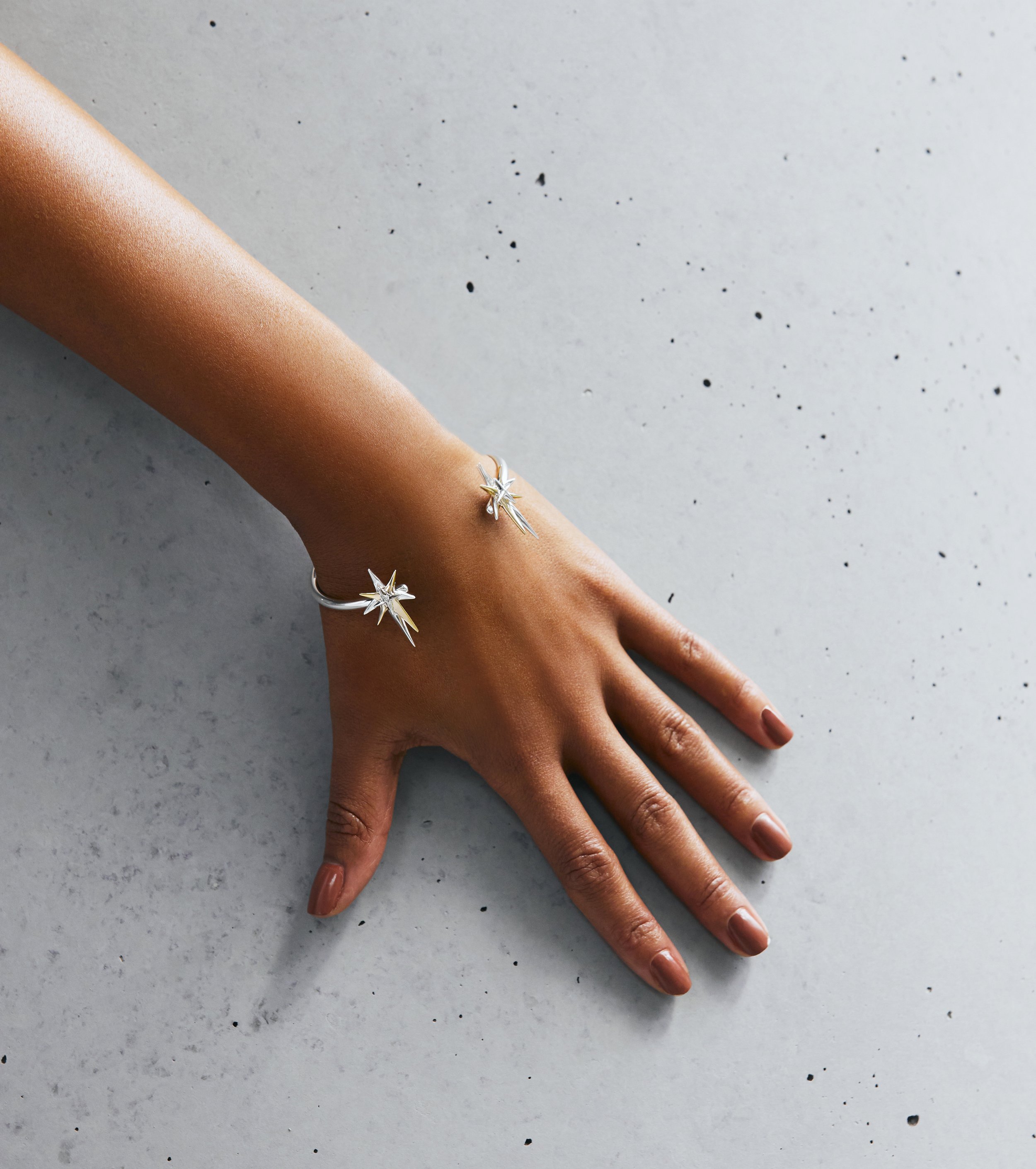 A hand with brown nail polish wearing two silver rings with star-shaped designs, resting on a gray speckled surface.