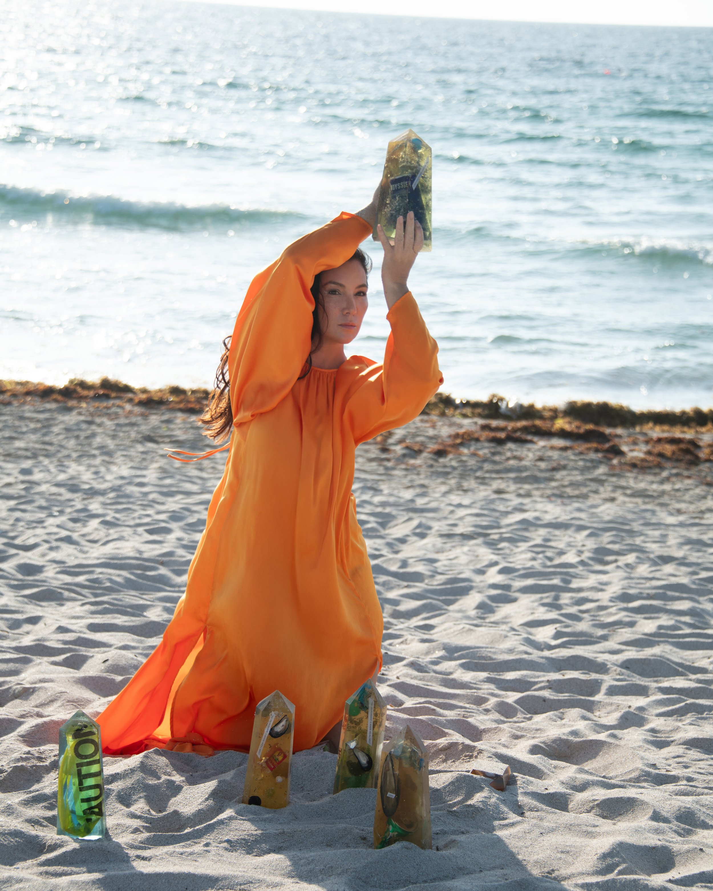 A woman in an orange dress kneels on a sandy beach near the ocean, holding a large green and clear crystal over her head, surrounded by smaller crystals and rocks.