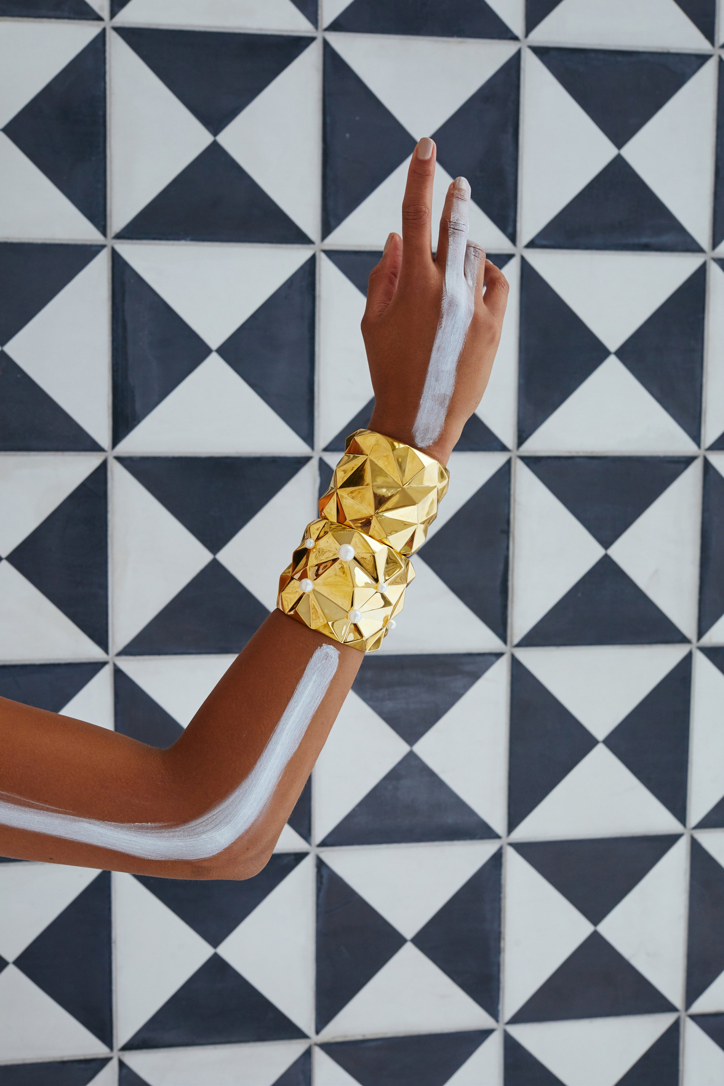 A person with dark skin tone wearing a large, gold, geometric bracelet with white accents, showing an arm painted with white lines, standing against a black-and-white tiled wall.