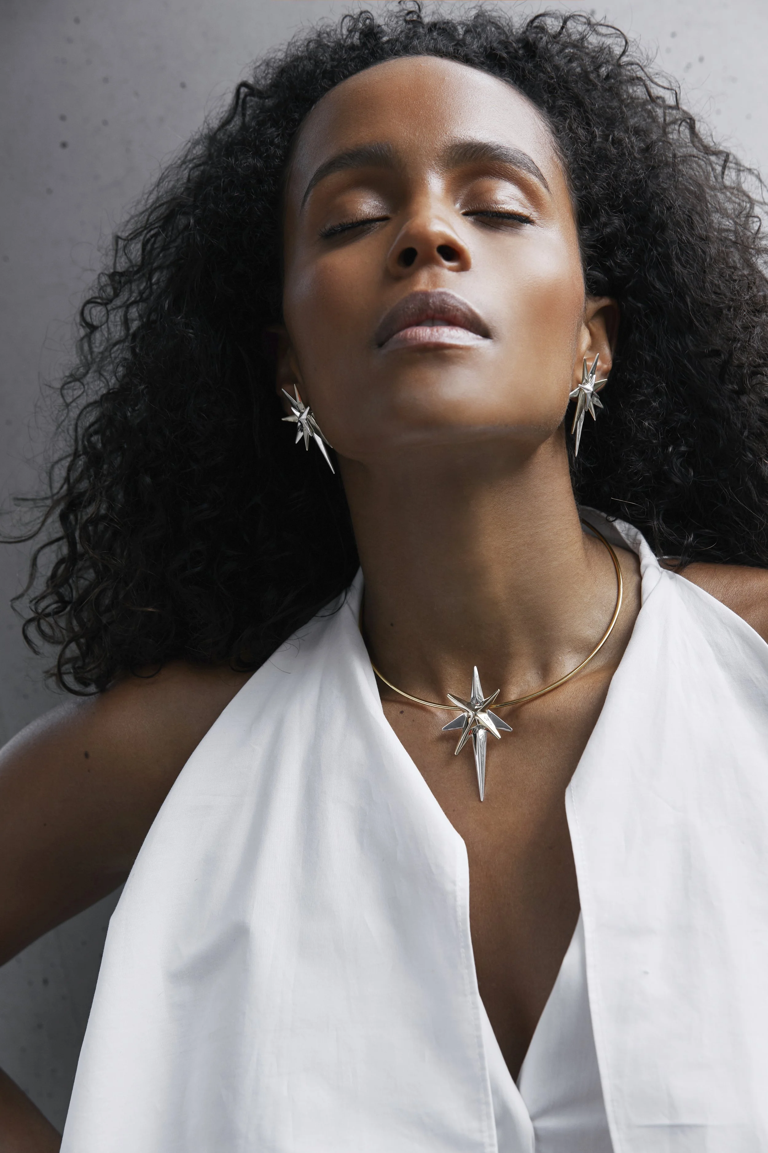 A woman with curly hair wearing silver star-shaped jewelry, a necklace, earrings, and a white sleeveless top, with her eyes closed.