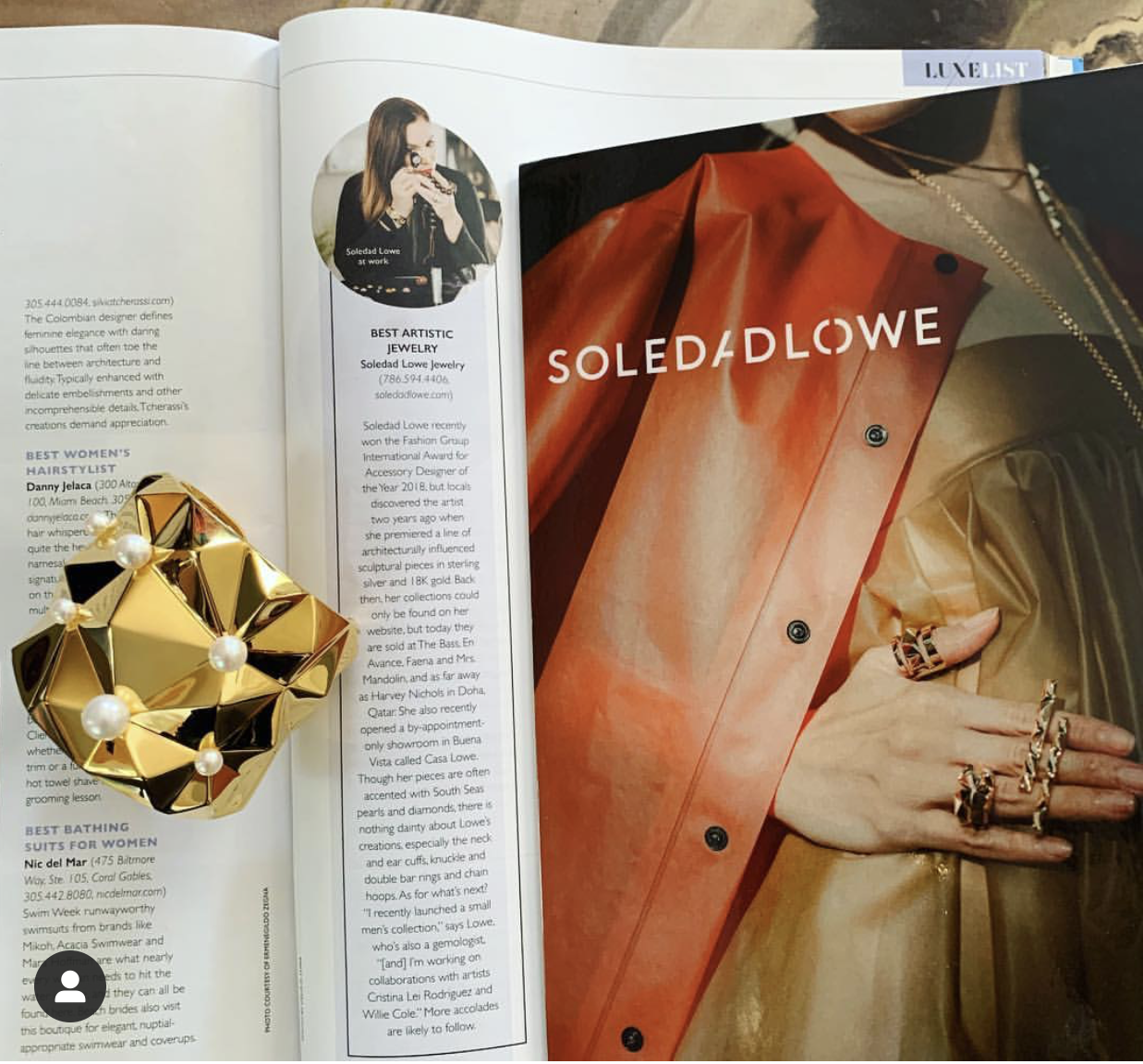 An open magazine showing a fashion spread with a model's hand adorned with multiple rings, a red and gold jacket, and jewelry. A gold geometric ring with pearl accents is placed on the magazine's pages.