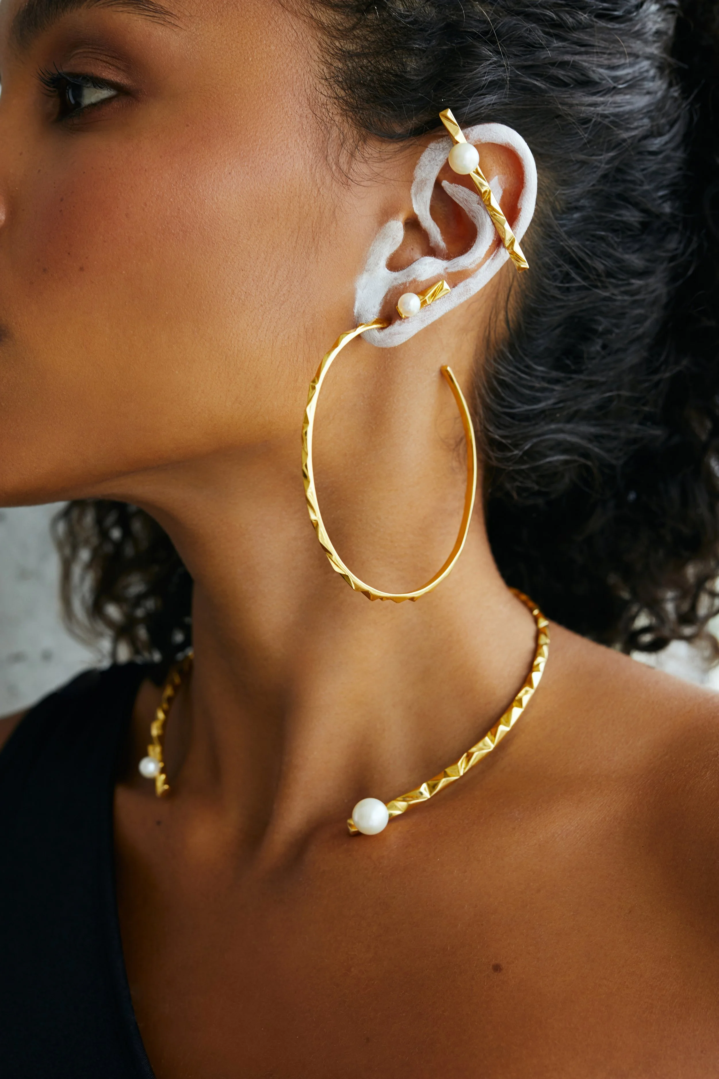 A woman with dark curly hair wearing multiple gold earrings and necklaces, decorated with pearls, with white paint outlining her ear.