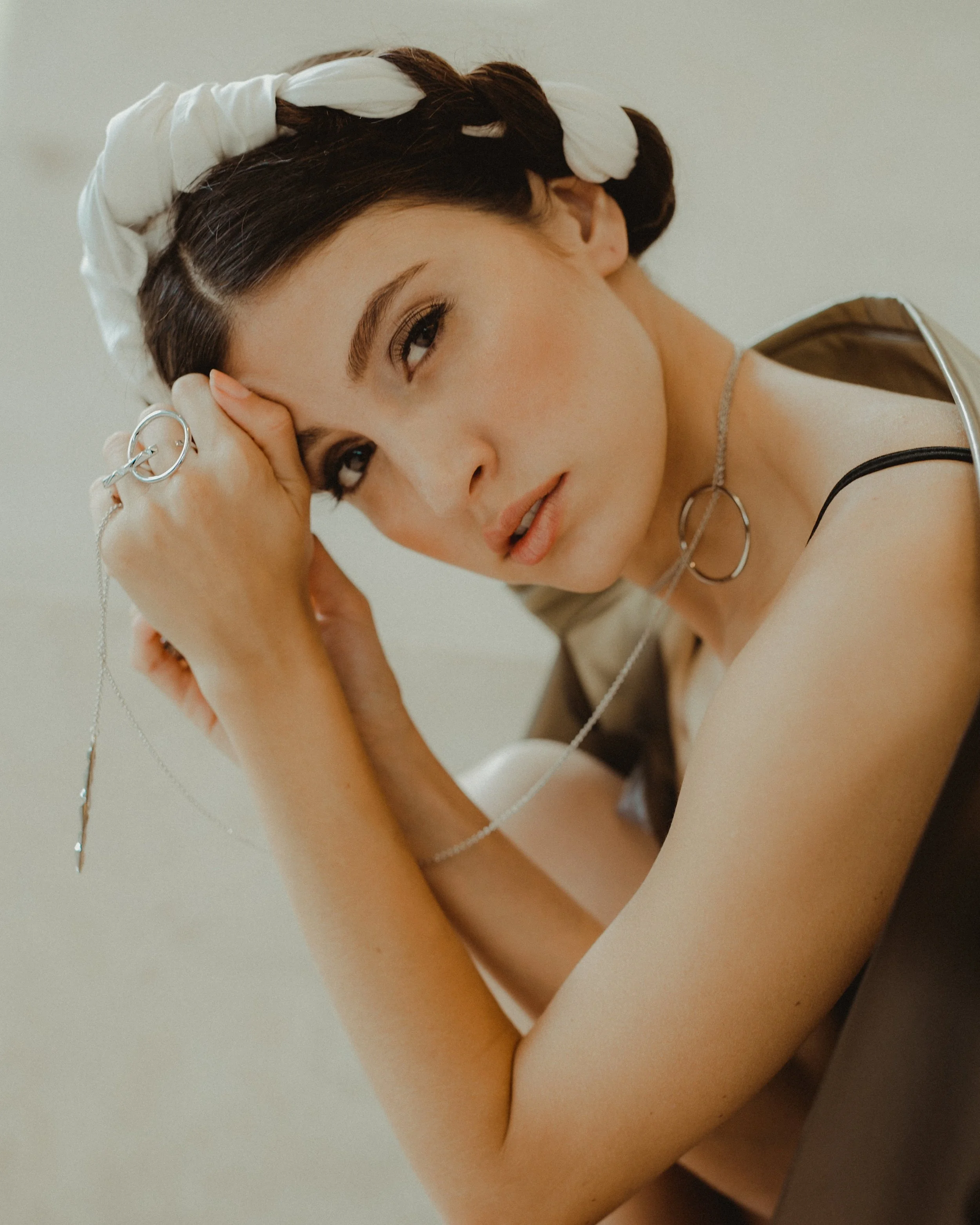 A young woman with dark hair in braids, wearing a white headband and silver jewelry, resting her head on her hand with a contemplative expression.