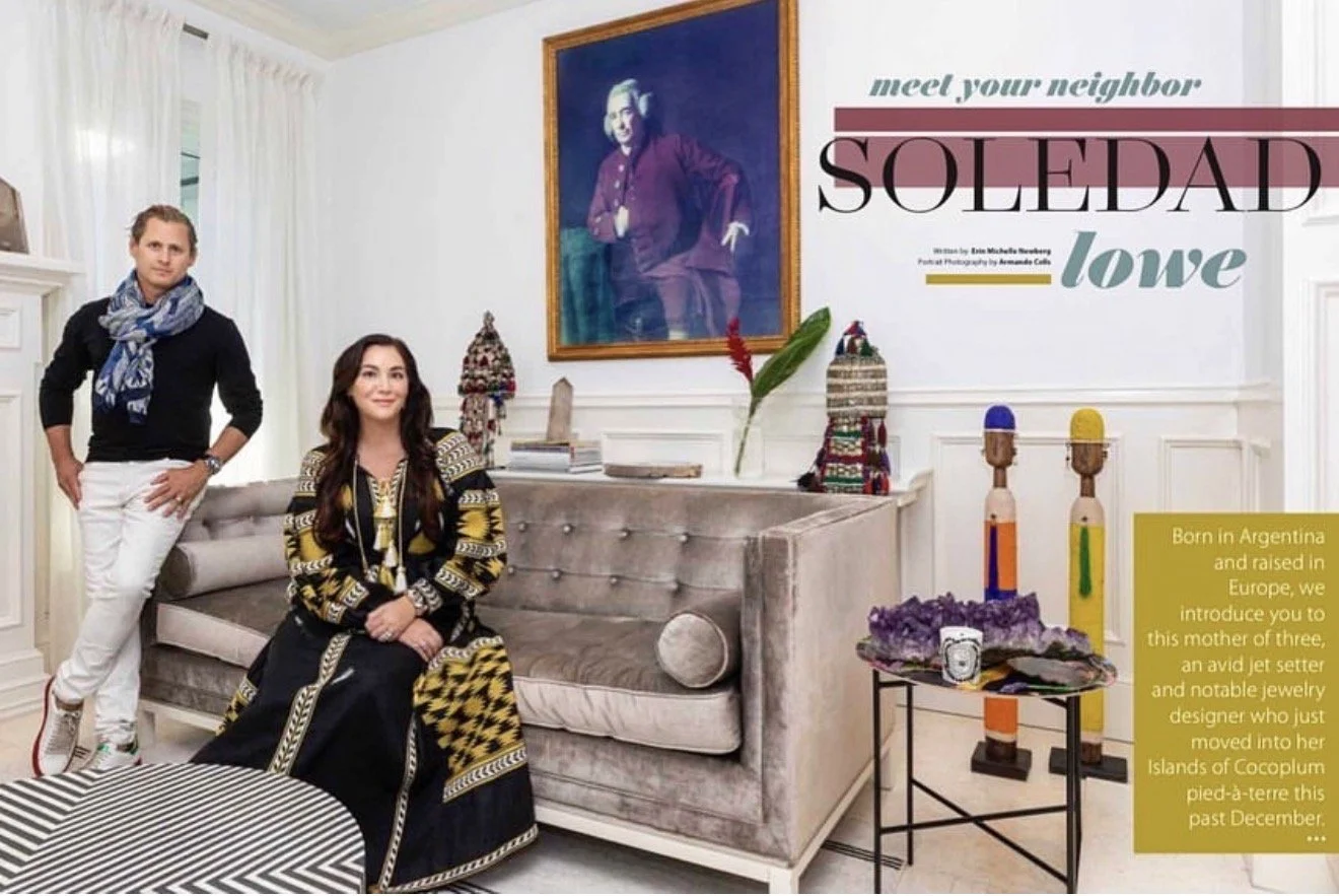 A woman seated on a beige sofa and a man standing next to her in a well-decorated living room with a large portrait of a woman on the wall, colorful decorations, and a small table with crystal and stone objects.