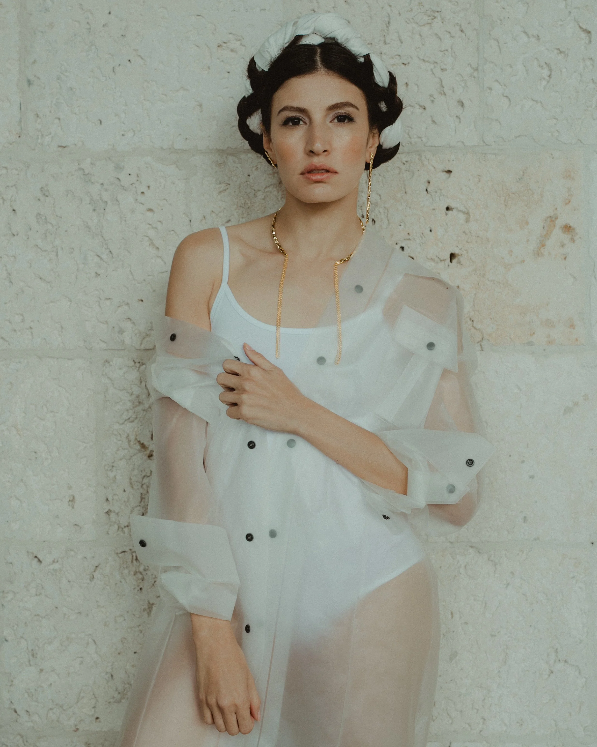 A woman with dark hair styled with a white braided headband, wearing a white tank top, a sheer white jacket with black buttons, gold jewelry, and posing against a textured white brick wall.
