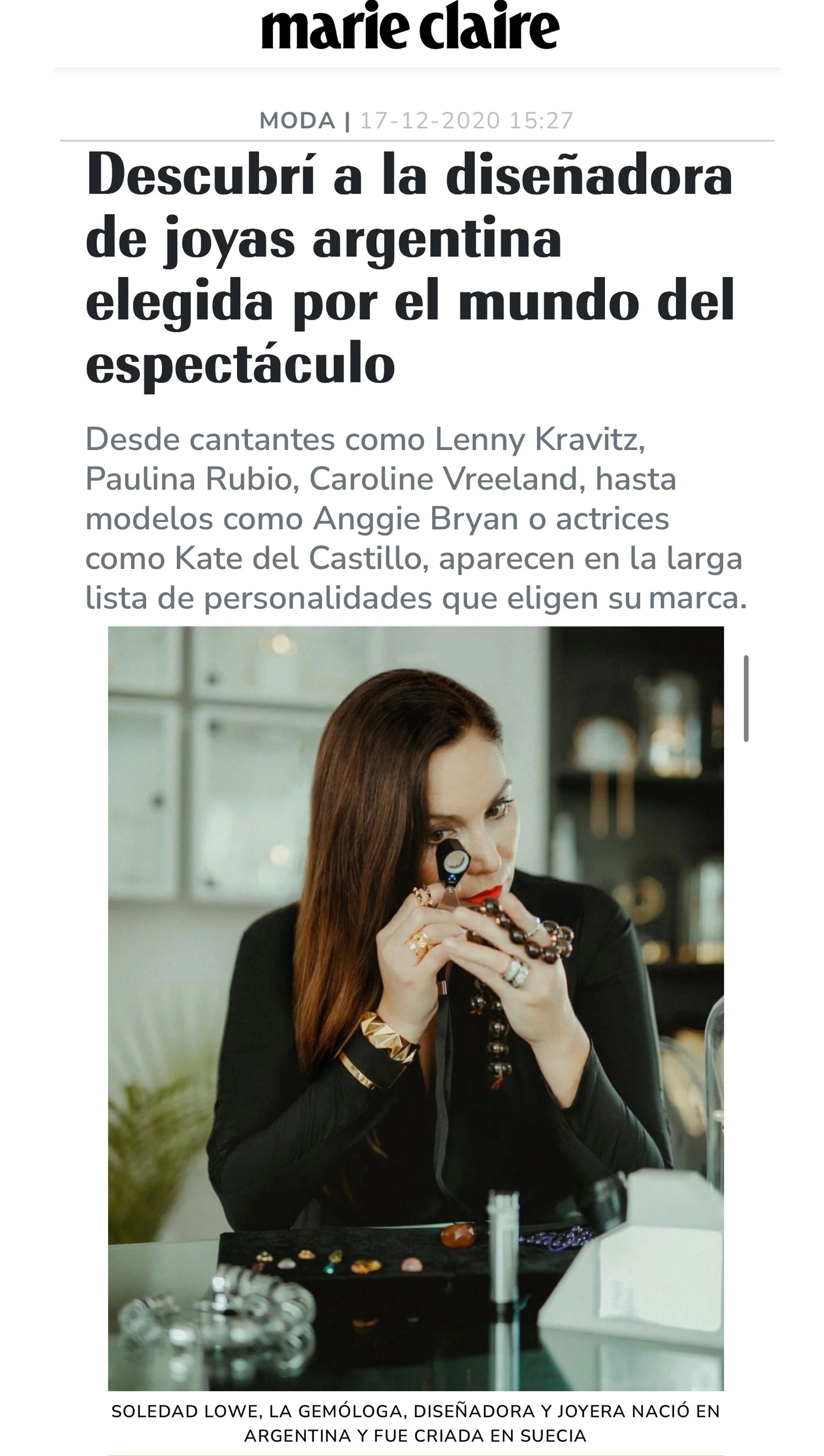 A woman with long brown hair, wearing black, holds a necklace close to her face while sitting at a table with jewelry in front of her. The background suggests an indoor setting.