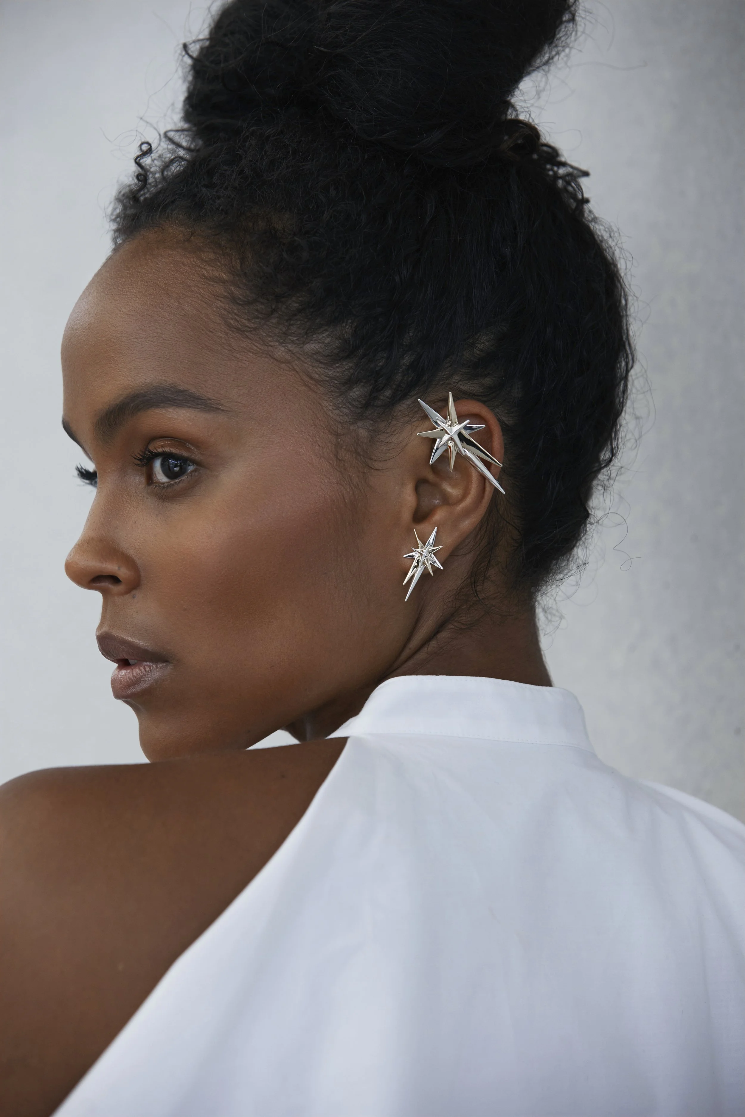A woman with dark skin and textured curly hair styled in a high bun, wearing large silver star-shaped earrings with multiple pointed spikes, and a white sleeveless garment against a plain gray background.