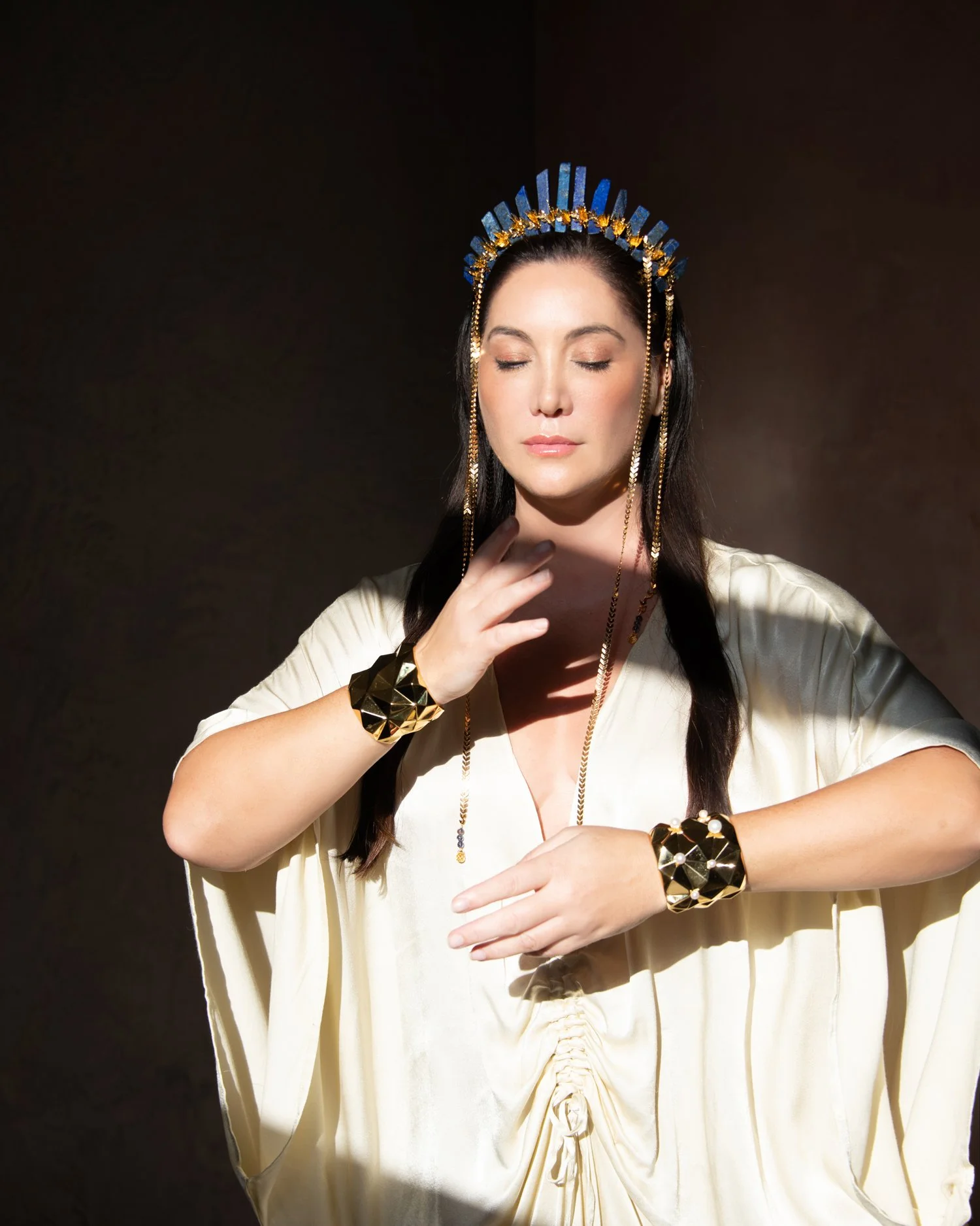 A woman with long dark hair wearing a cream-colored dress and gold jewelry, including large bracelets and a necklace, with her eyes closed and one hand near her face, standing against a dark background with dramatic lighting.