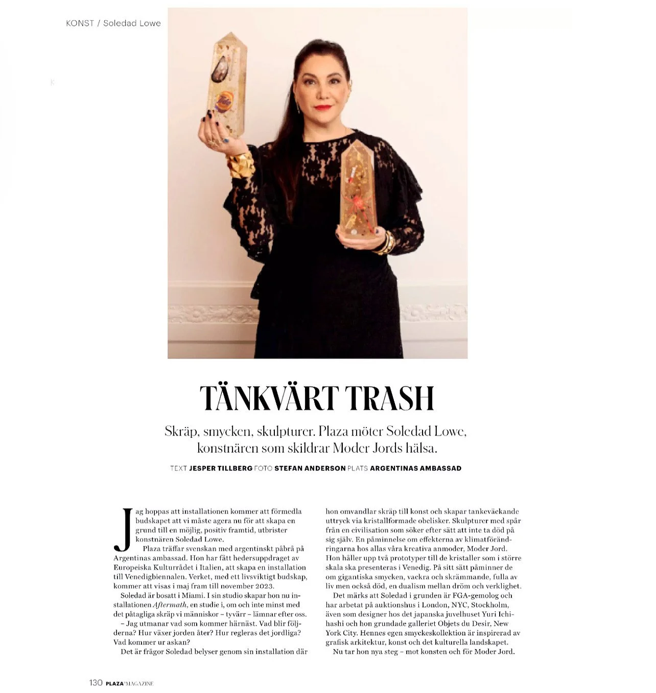 A woman in a black lace dress holding two wooden sculptures. The background is plain and light-colored, with some jewelry and jewelry on her wrists.