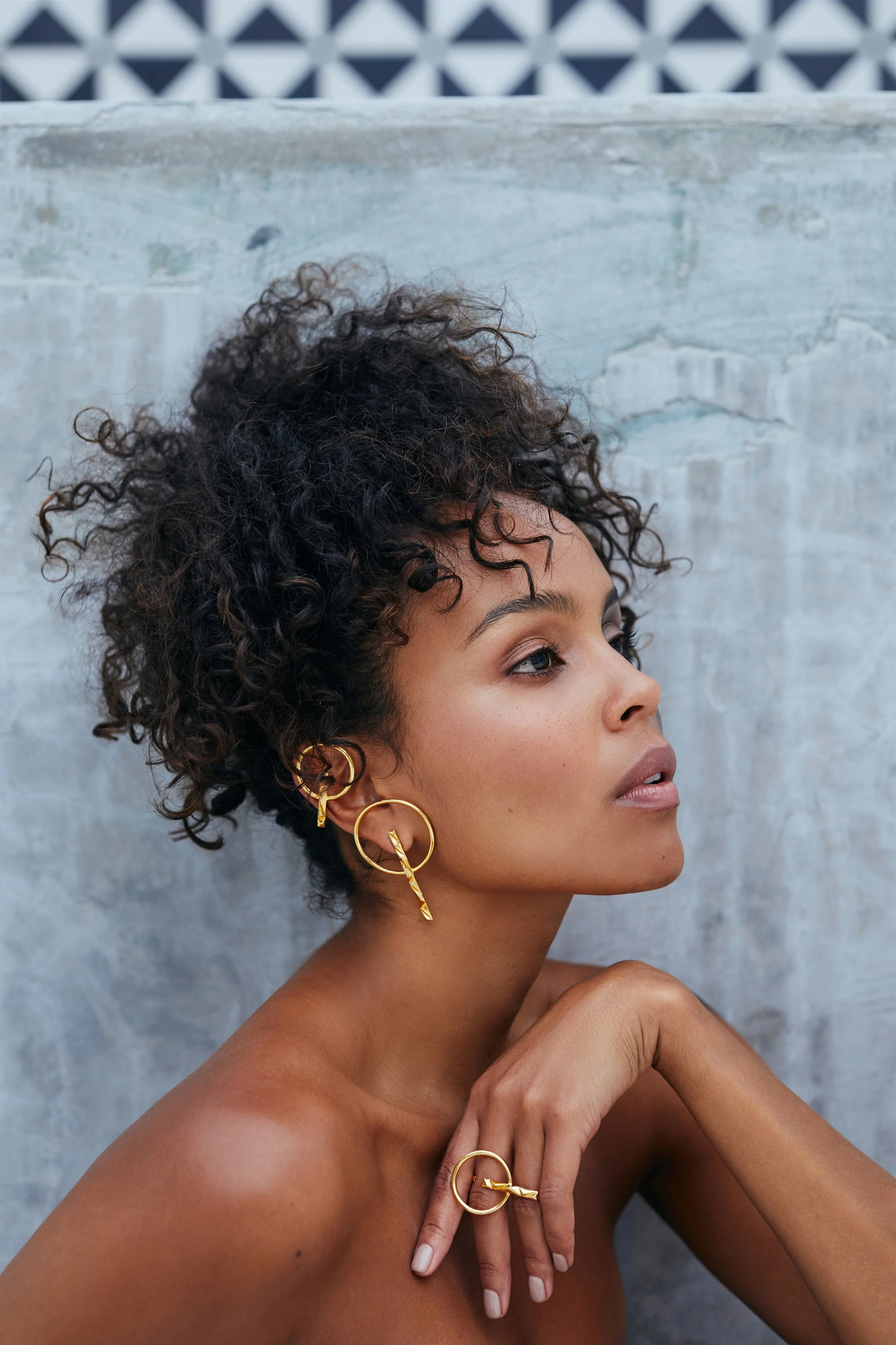 A woman with curly short hair wearing gold jewelry, including earrings, rings, and a necklace, posing against a concrete wall.