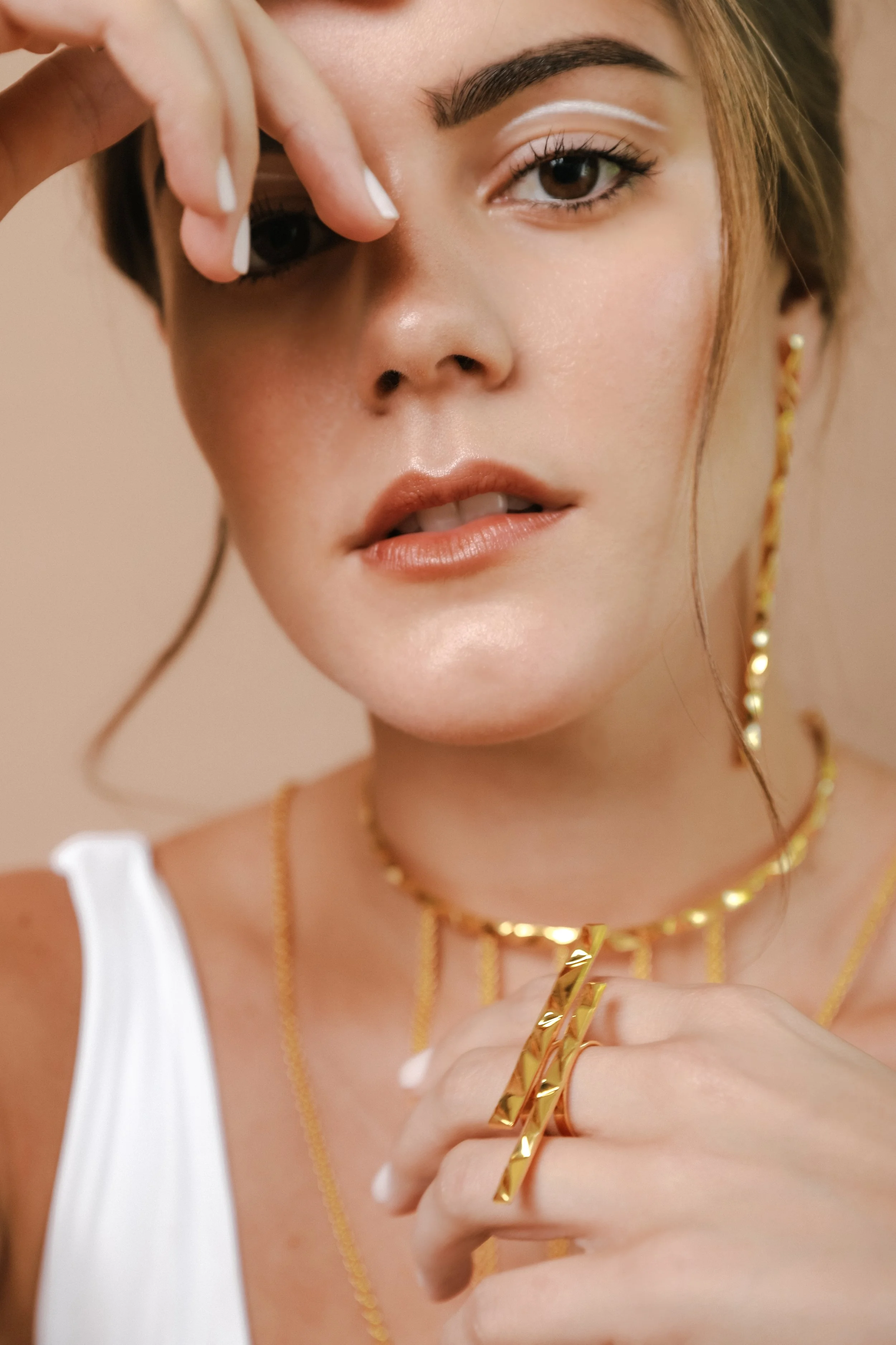 A woman wearing gold jewelry, including a necklace, earrings, and rings, with her hand near her face. She has makeup with white eyeliner and nude lipstick, and is wearing a white top.