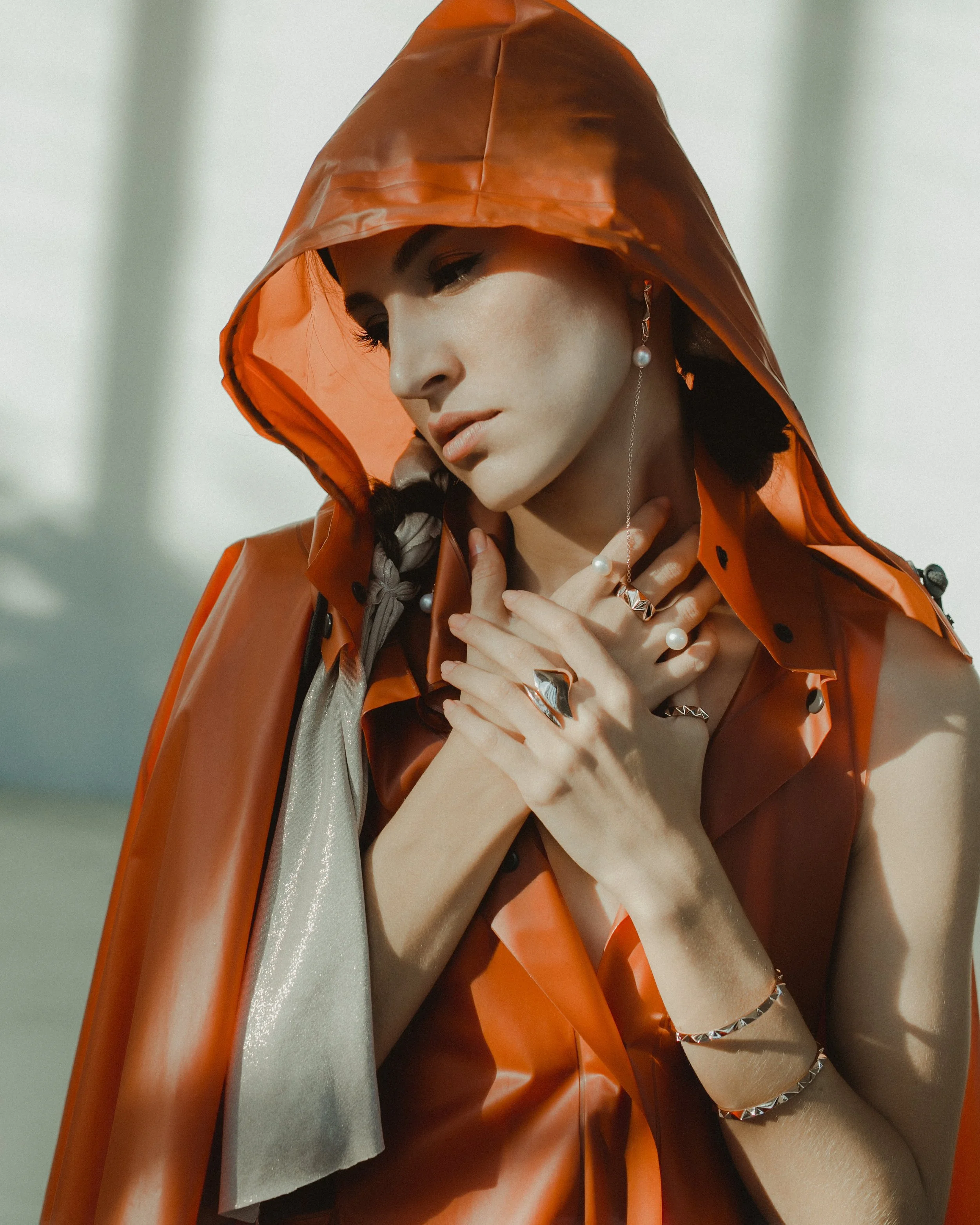 A woman wearing a shiny orange raincoat with the hood up, posing with her hands near her neck, adorned with various jewelry, in a bright setting.