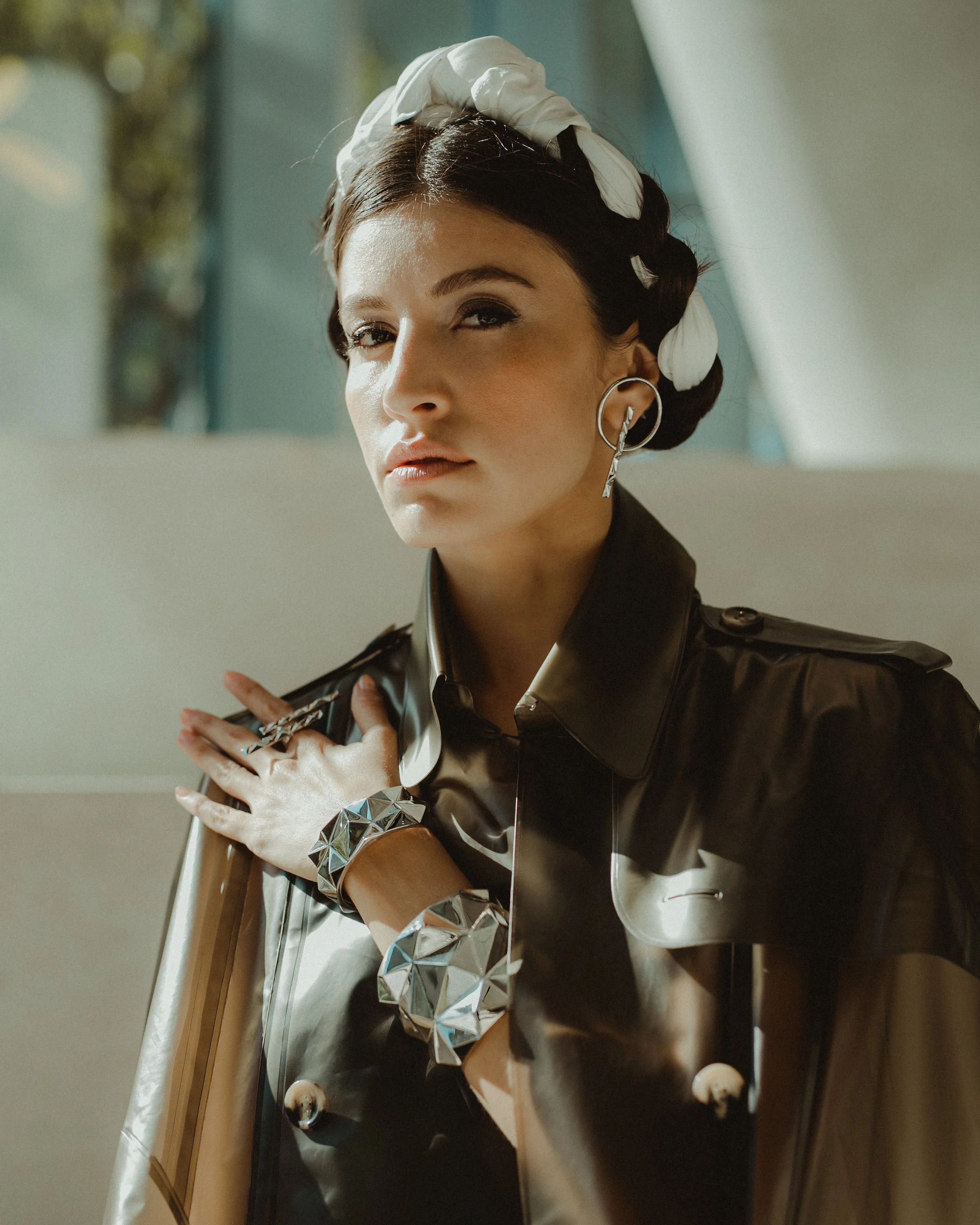 A woman with dark hair styled in an updo, wearing a white headband with feathers, large hoop earrings, a reflective metallic bracelet, and a shiny black trench coat, posing indoors with one hand on her chest.