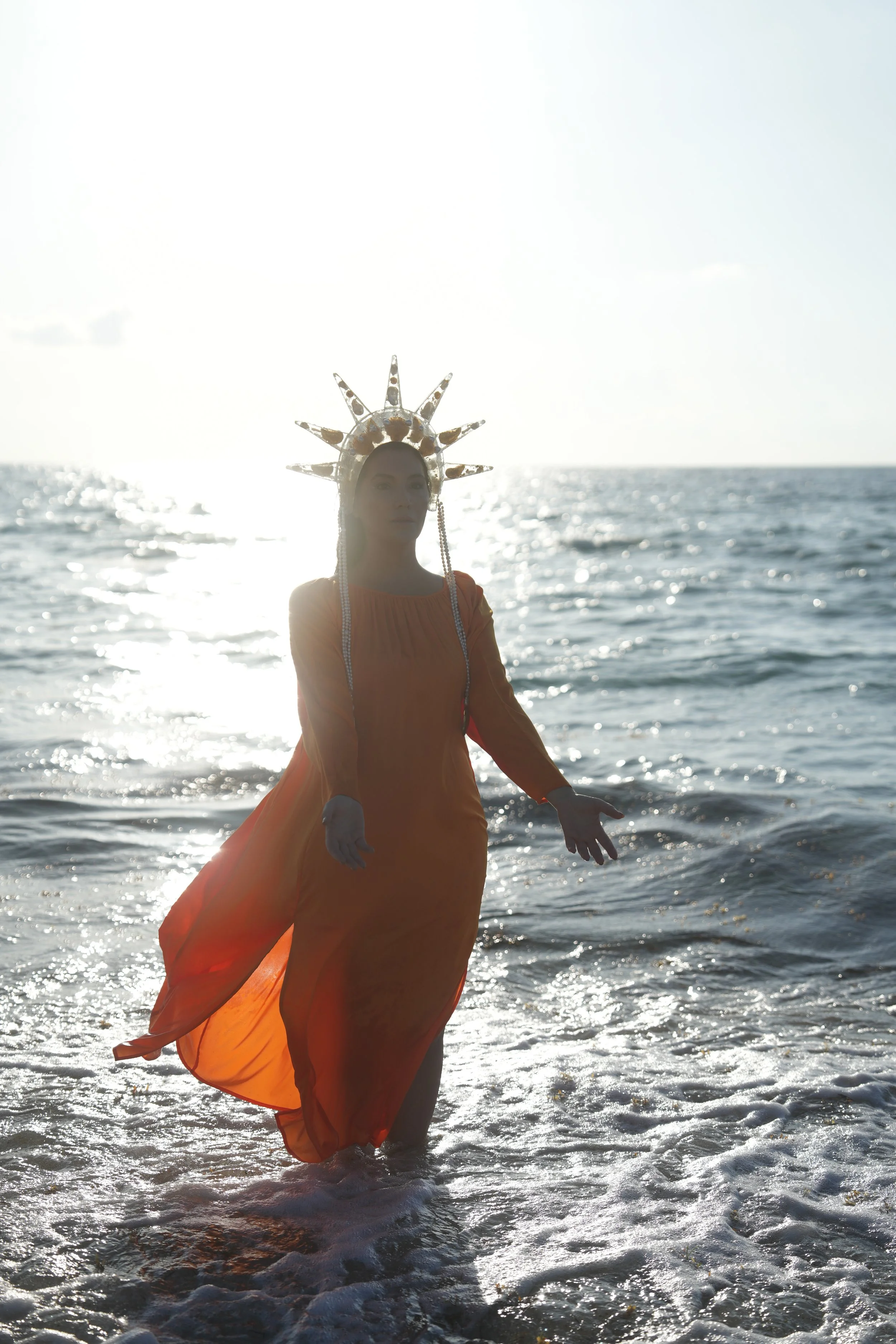 Person dressed as a religious or mythical figure in an orange dress with a crown, standing in the water at the beach during sunset.