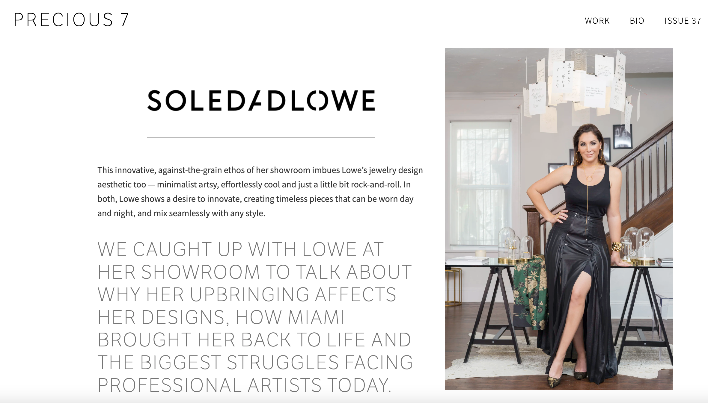 Woman standing in a room with a flight of stairs behind her, wearing a black tank top and a long black skirt with a high slit. The room has a window, a table with glass cloches, and artwork on the walls.