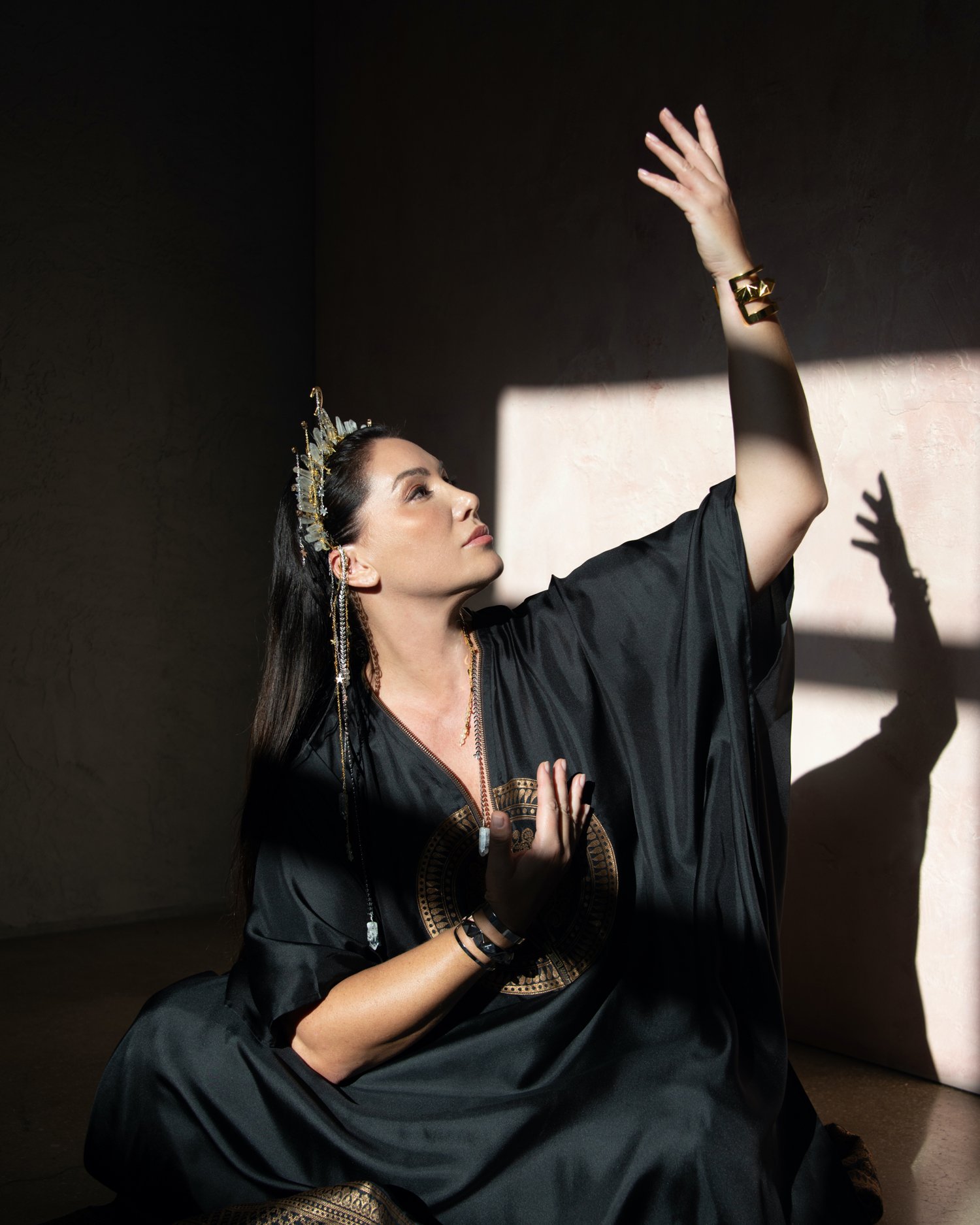 A woman dressed in black, wearing a crown and jewelry, sitting on the floor with a dramatic light and shadow effect, reaching up with her right hand.