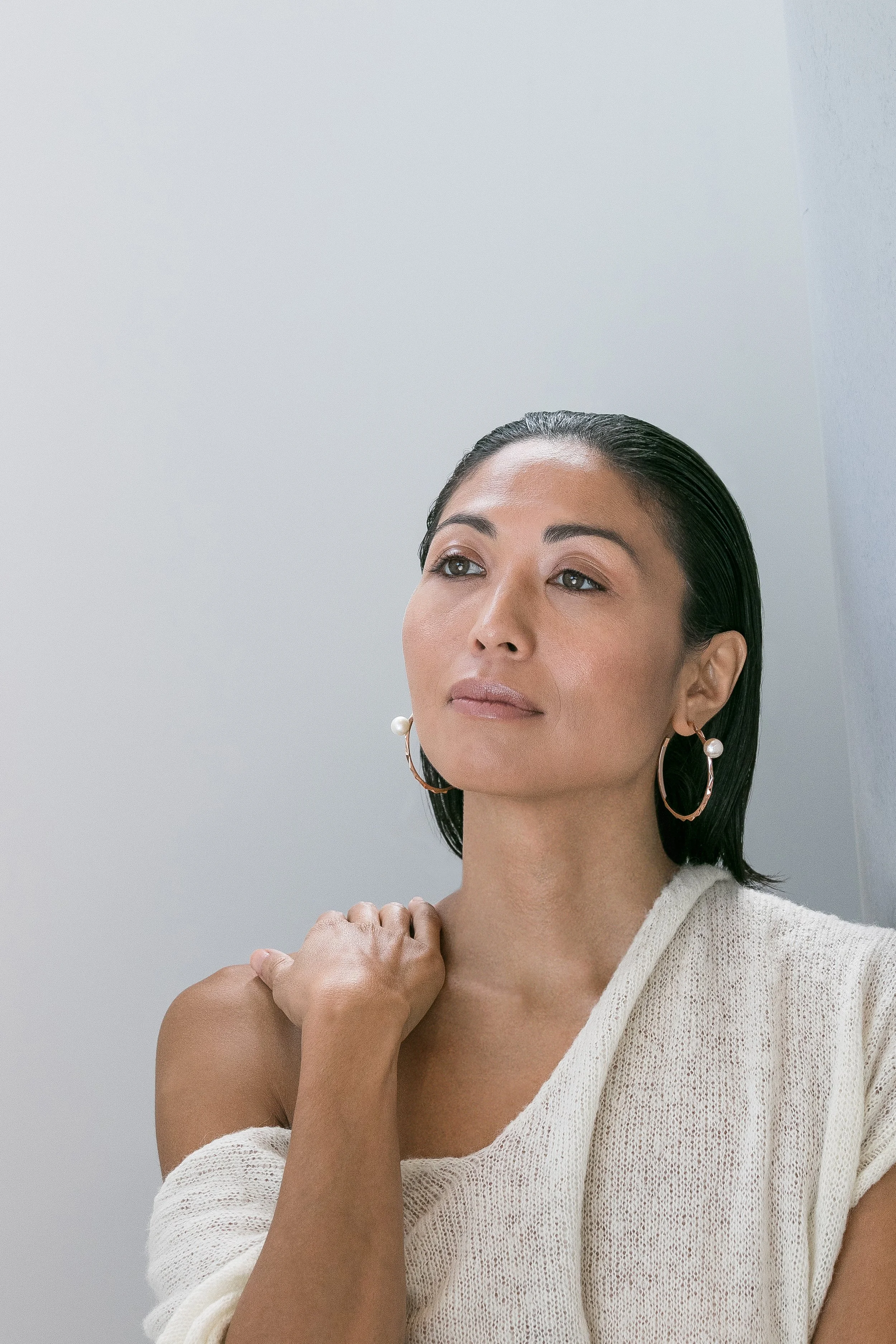 A woman with dark hair, wearing large hoop earrings with pearls and a cream-colored sweater, looking contemplative with her hand on her shoulder.