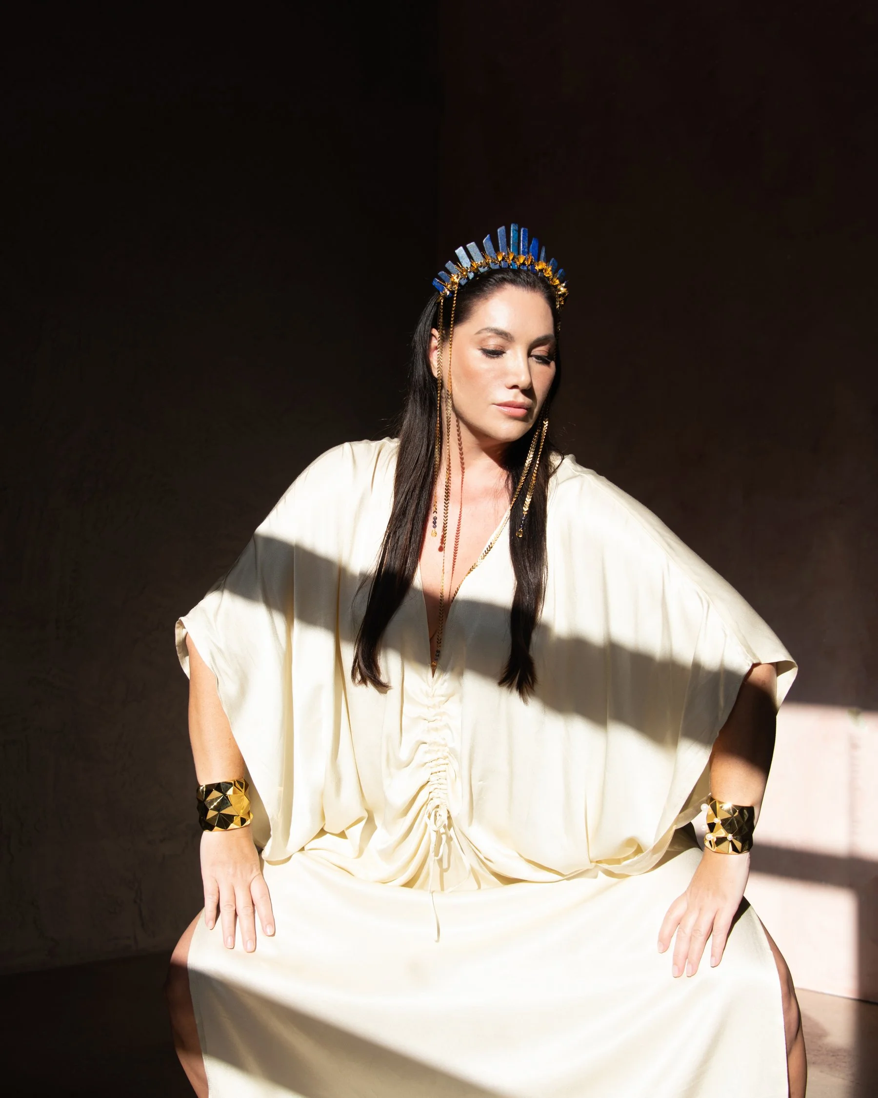Woman wearing a gold dress, gold jewelry, and a blue and gold headpiece, sitting with her eyes closed in sunlight.