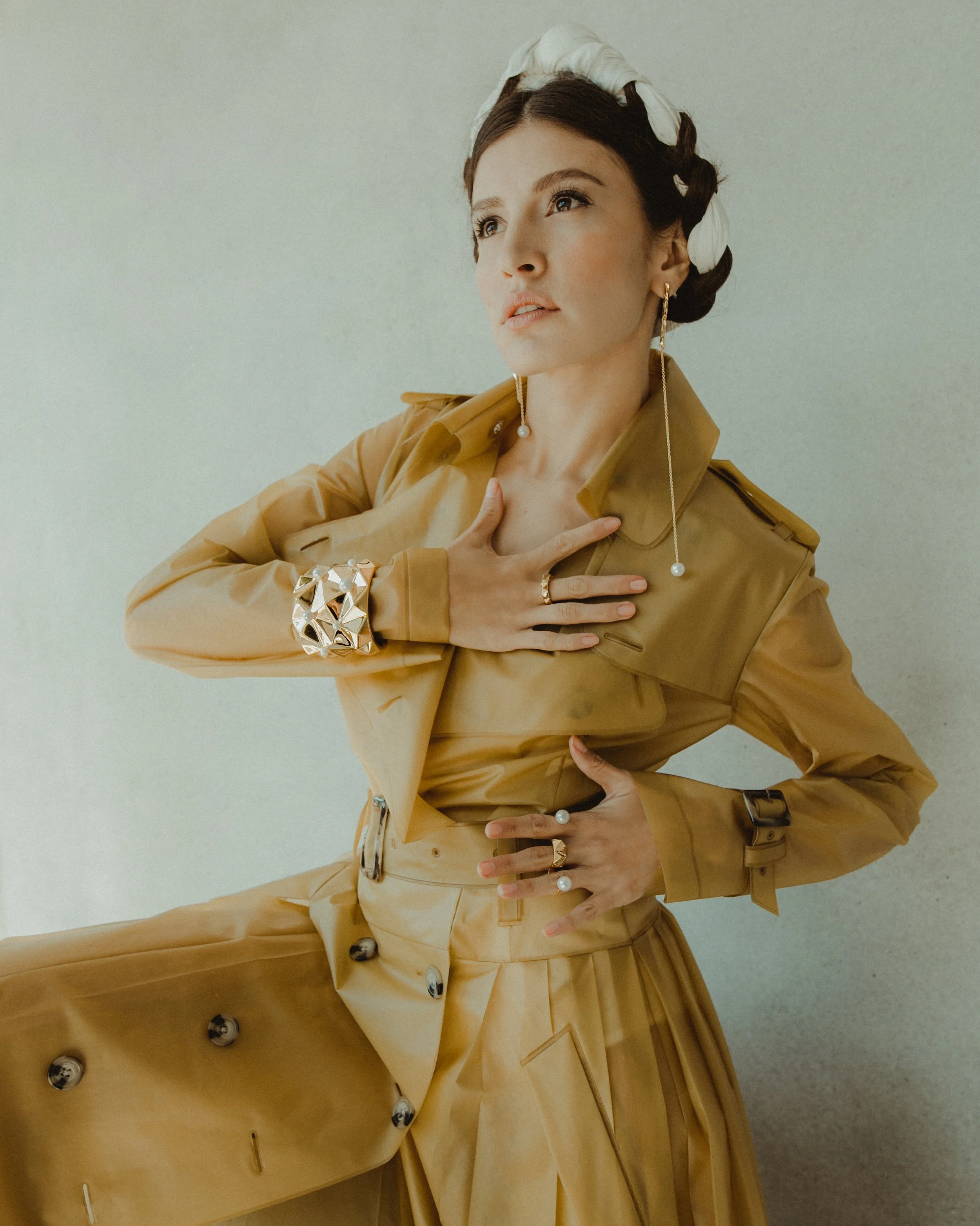 A woman with dark hair styled in braid with a white cloth head wrap, wearing a khaki trench coat with belt and buttons, accessorized with pearl earrings, rings, a cuff bracelet, and a long pearl chain earring. She is posing with her hand on her chest