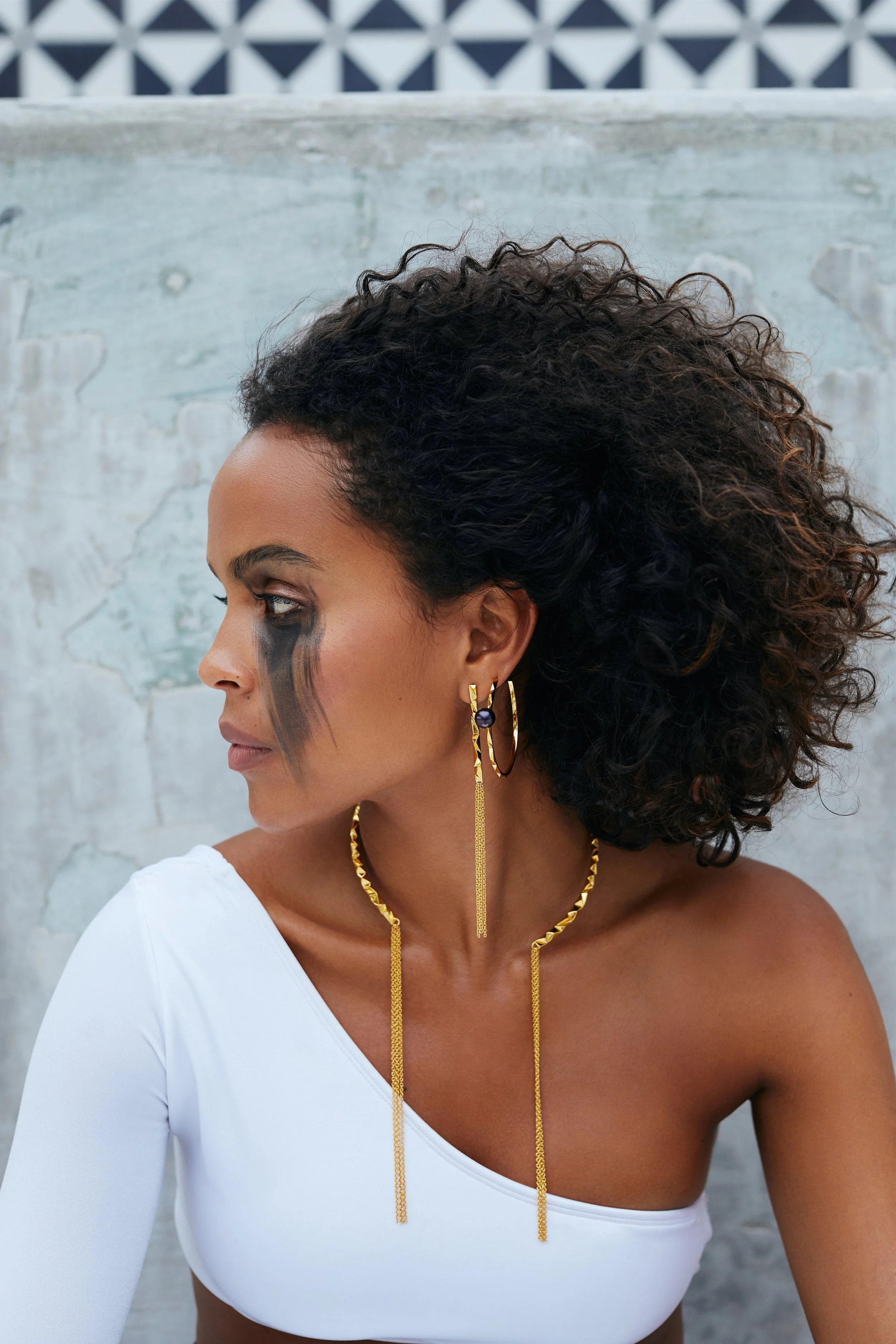 A woman with curly black hair and dark skin, wearing gold jewelry and a white one-shoulder top, has black makeup streaks under her eye, and stands against a textured concrete wall.